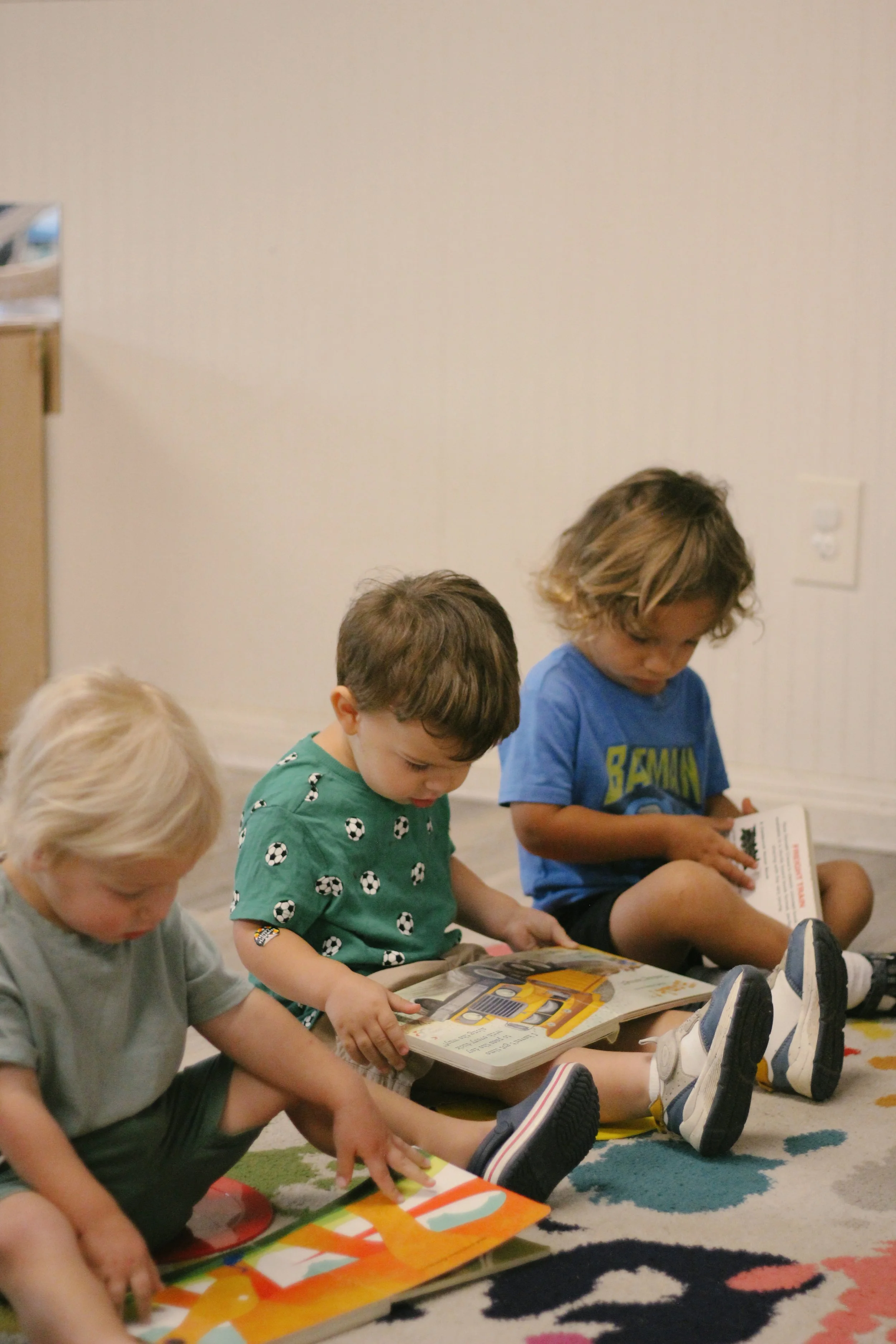Three young children sitting on a colorful rug, reading picture books.