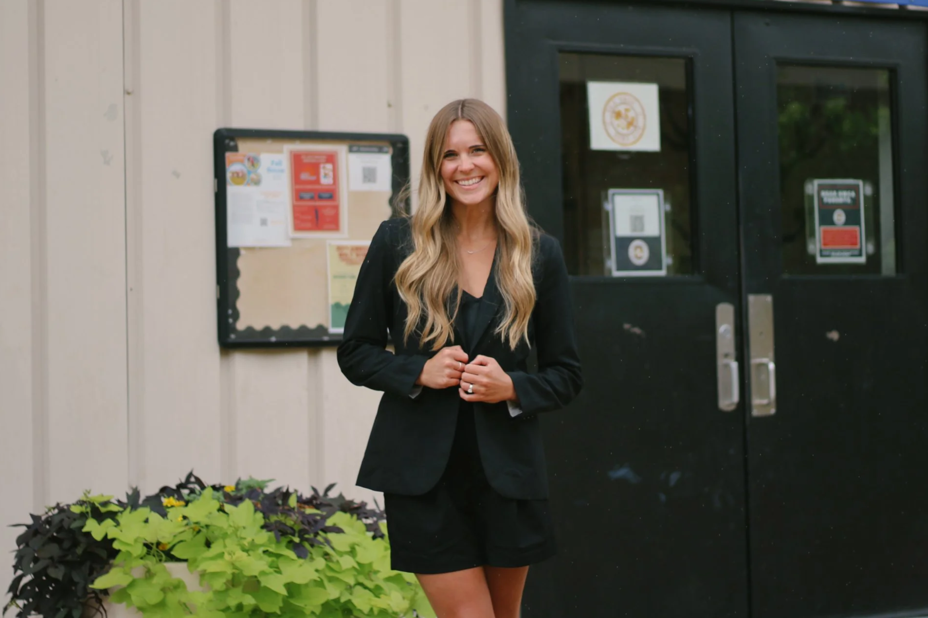 A smiling woman with long wavy blonde hair wearing a black blazer and shorts standing outside near black double doors and a notice board.