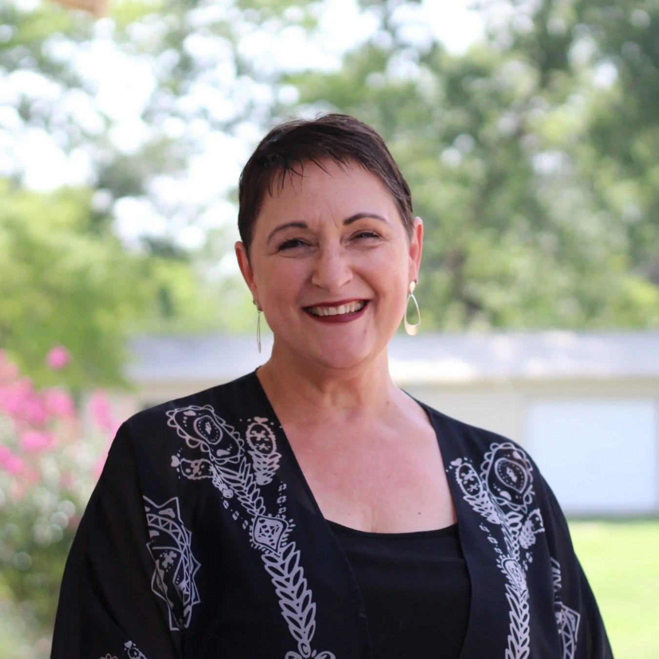 Smiling woman with short dark hair, wearing earrings and a black top with lace detail, standing outdoors with trees and pink flowering bushes in the background.