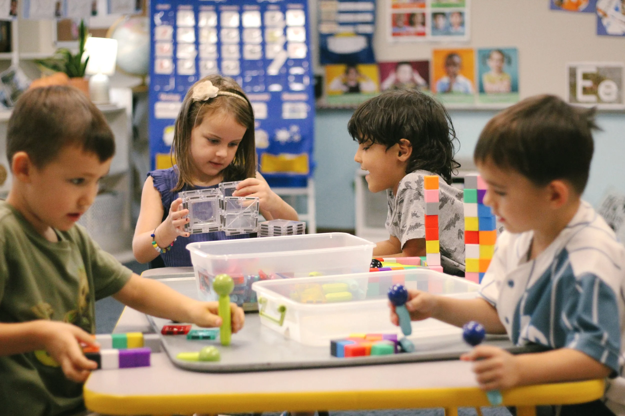 Four young children sit around a table in a classroom, playing with colorful building blocks, engaging in a group activity.