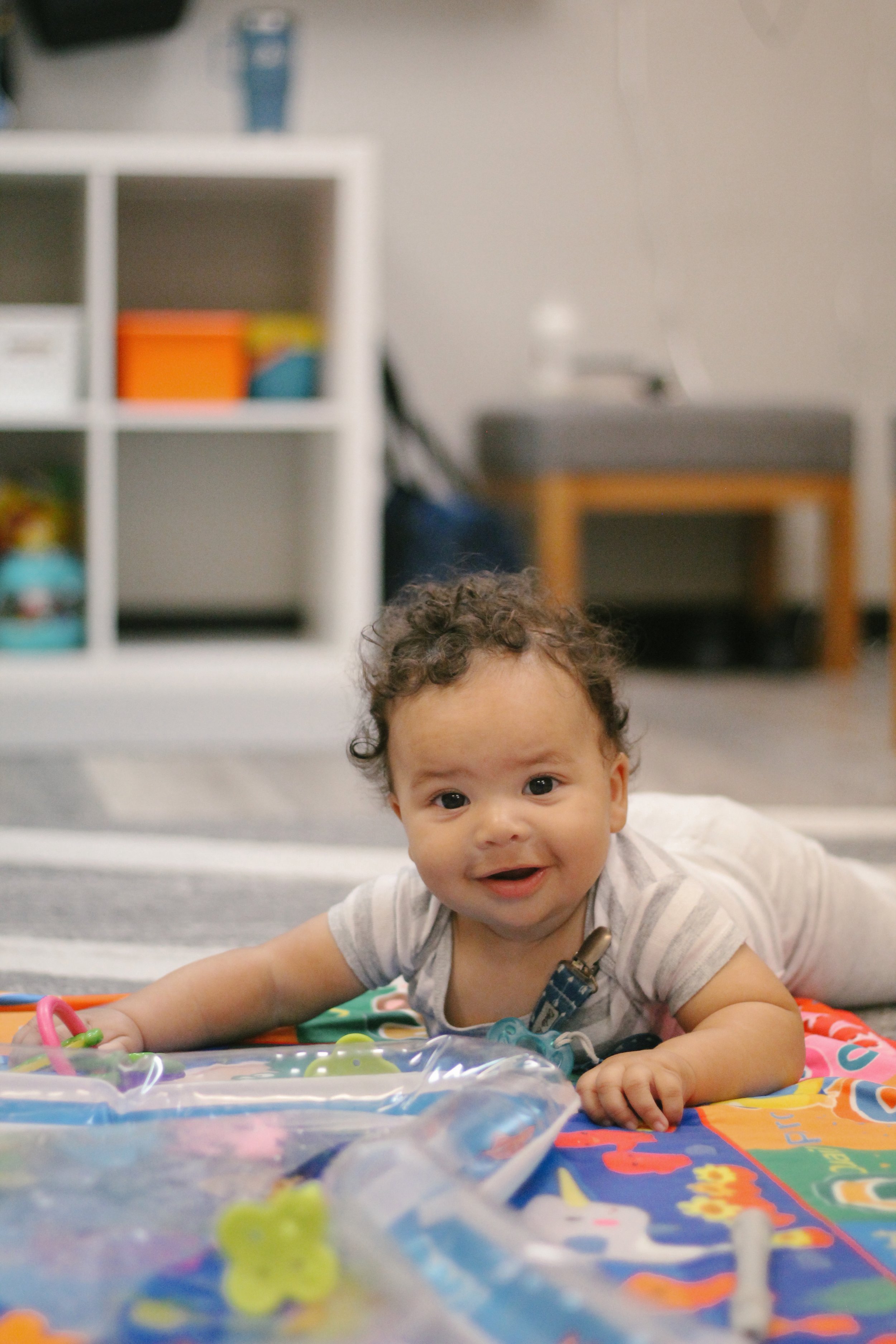 A baby lying on their stomach on a colorful play mat, smiling at the camera.