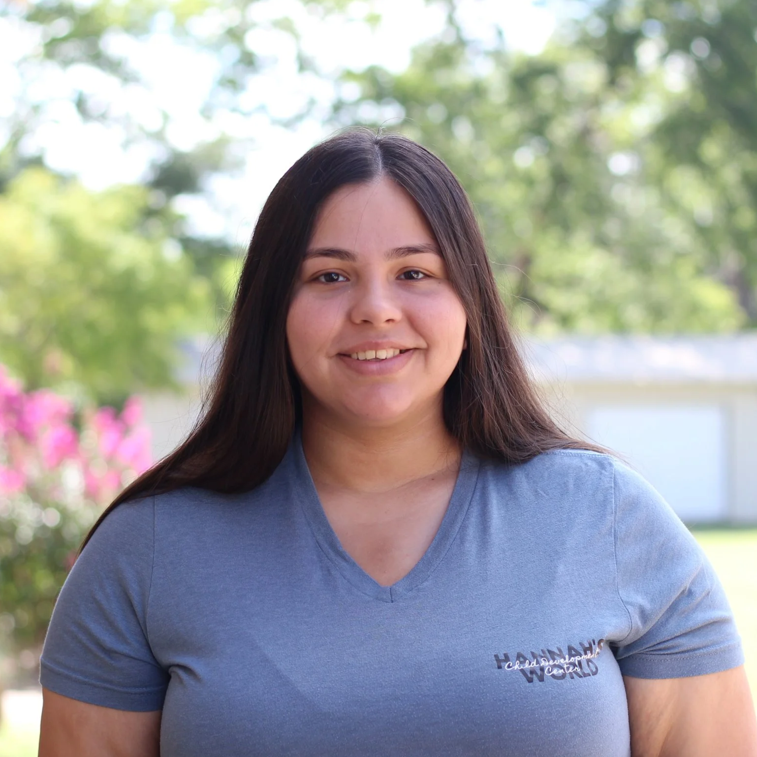 Portrait of a young woman with long dark hair, smiling, wearing a blue T-shirt, outdoors with greenery and pink flowers in the background.
