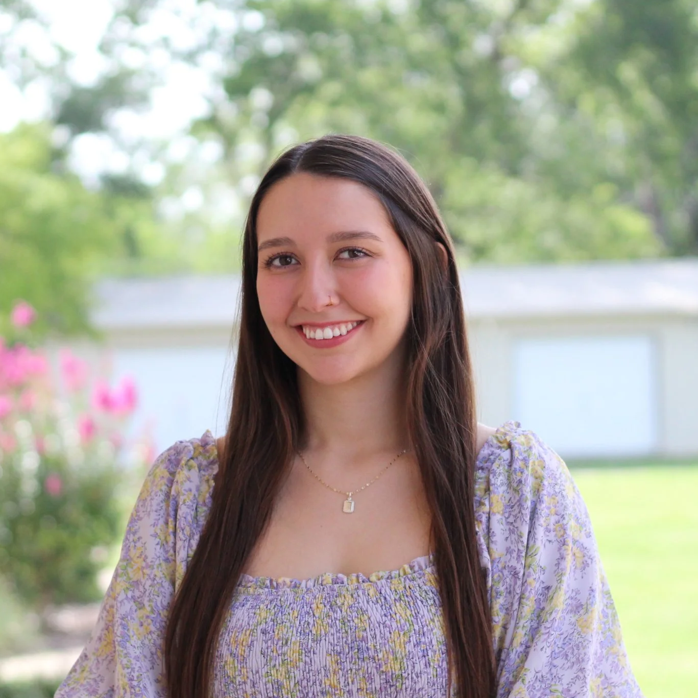 Young woman outdoors with long brown hair, smile, wearing a purple floral dress and a necklace, background of green trees and a white fence.