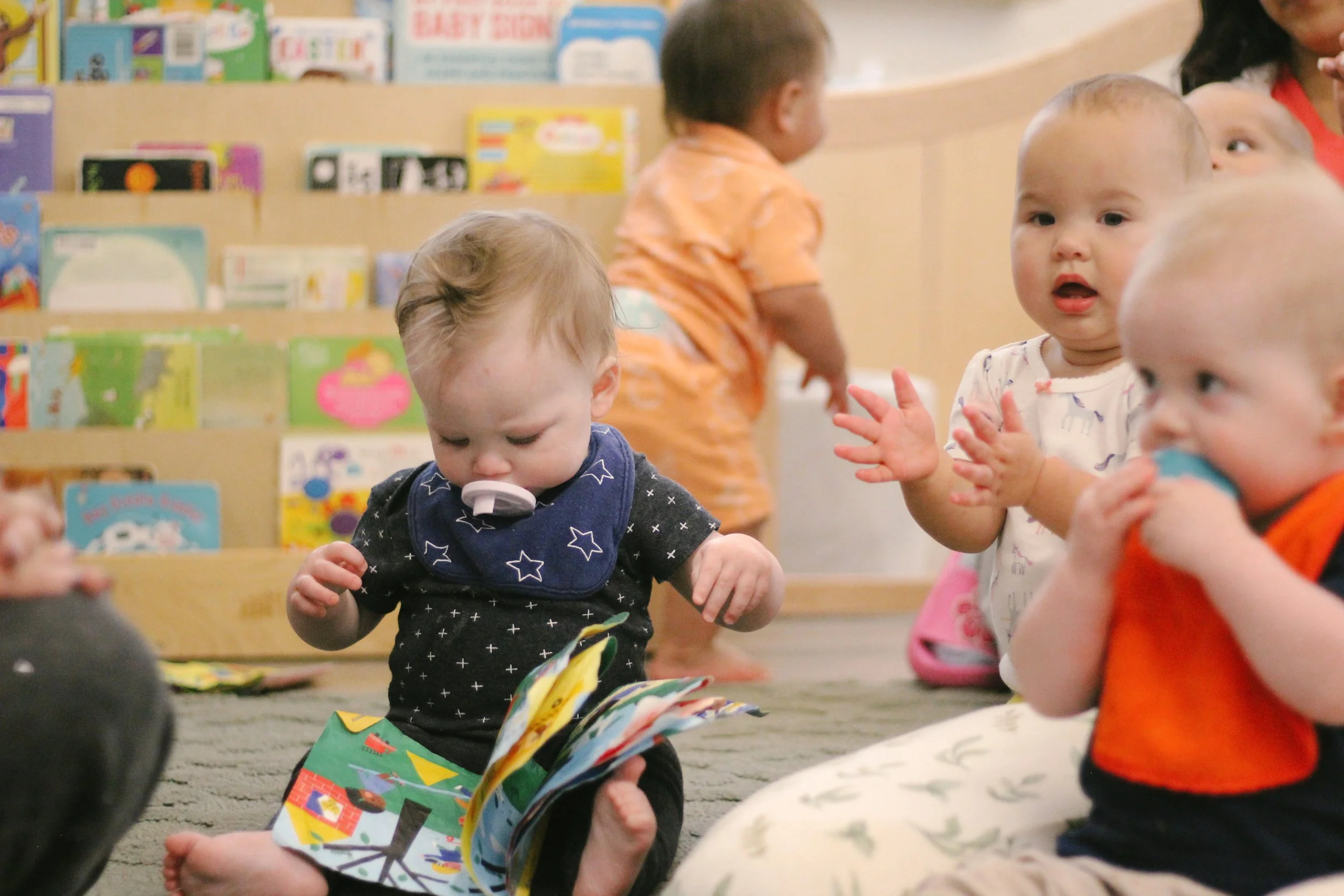 Infants in a classroom sitting on the floor; one child looking at a colorful book, others engaging and clapping, with shelves of books in the background.