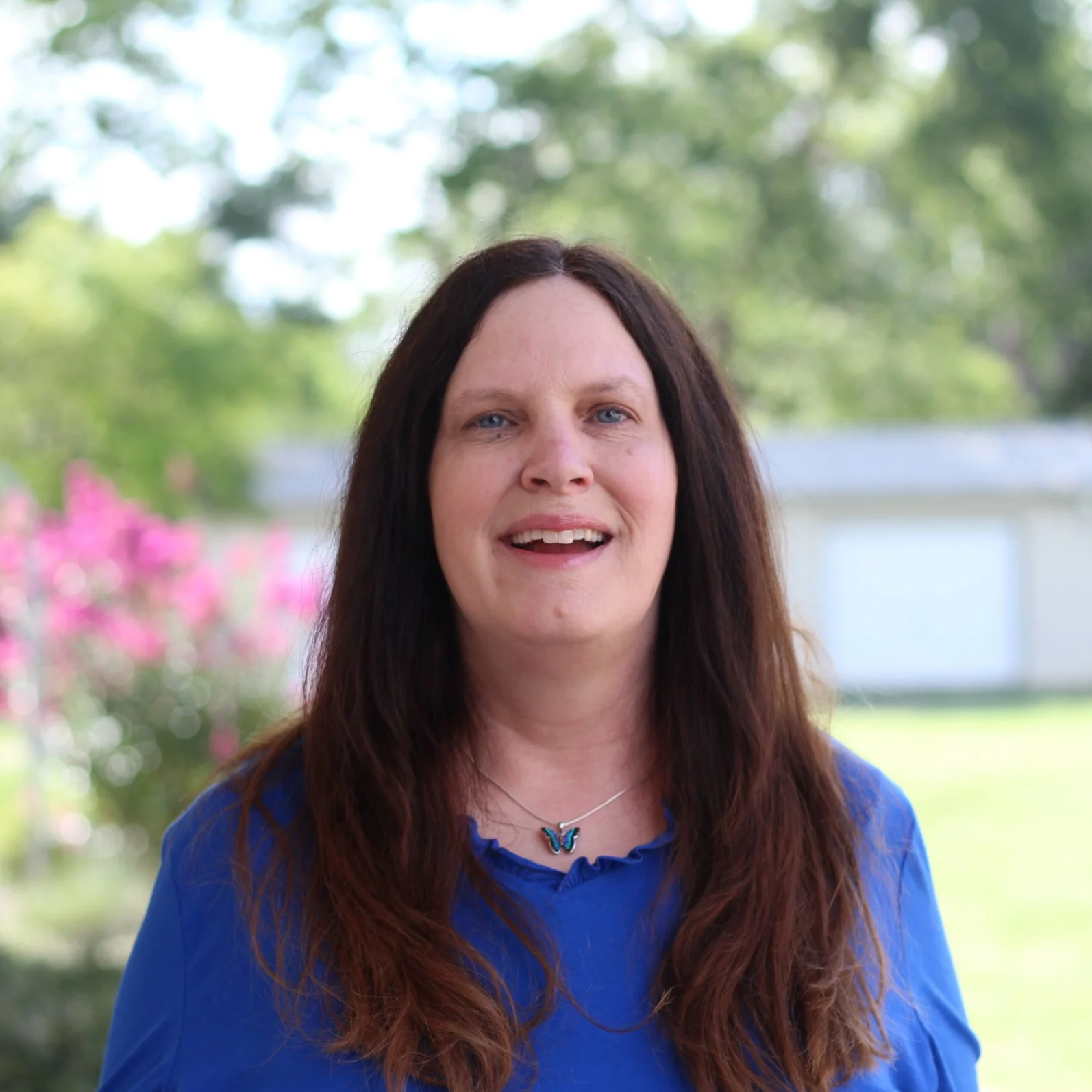 Portrait of a woman smiling outdoors with trees and pink flowering bushes in the background.