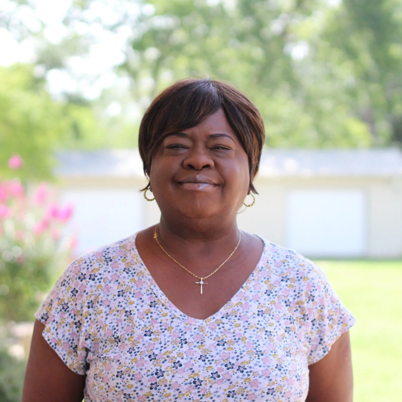 A woman stands outside in a garden with trees and bushes in the background, wearing a floral top and jewelry, with a slight smile on her face.
