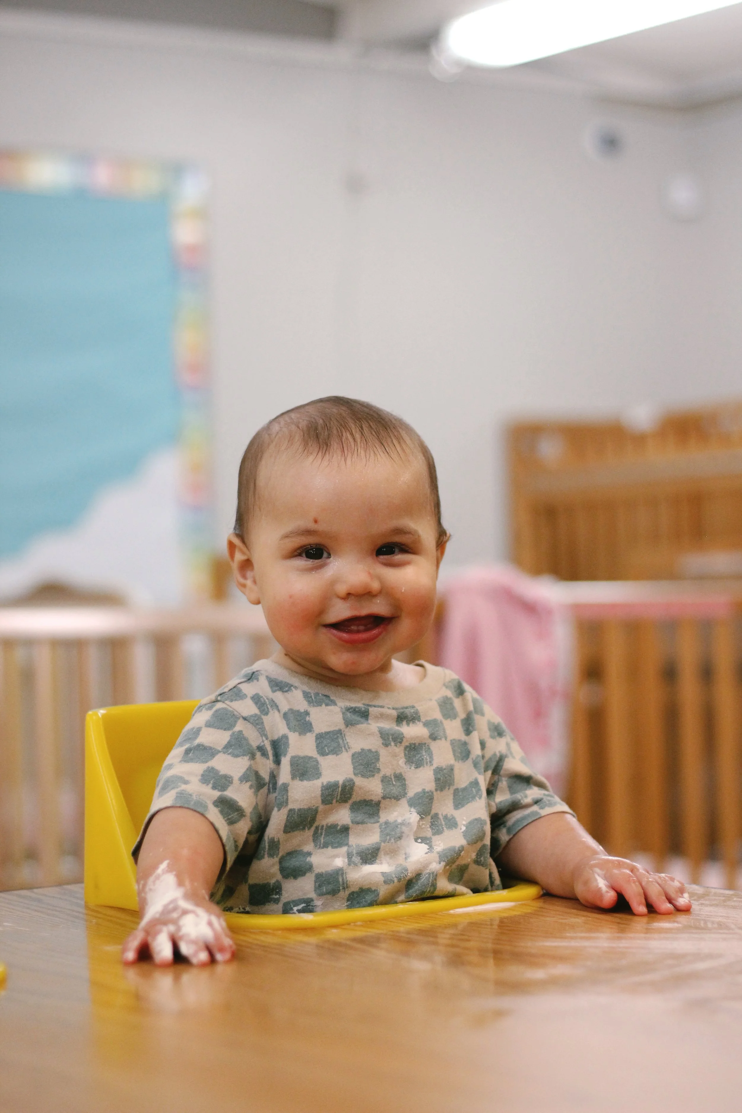 A happy toddler sitting at a table in a classroom, with messy hands and face.