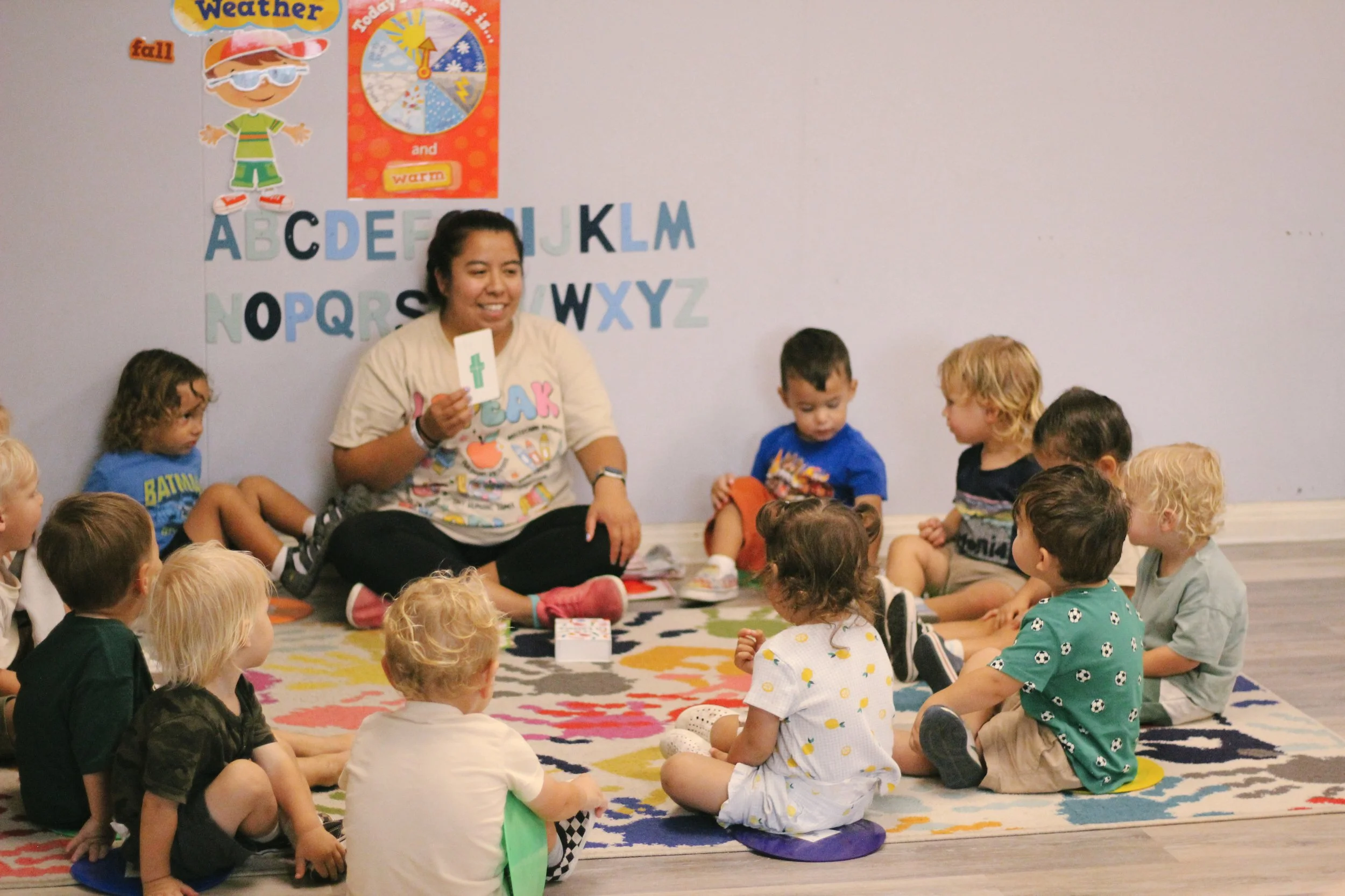 A group of young children sitting on a colorful rug in a classroom listening to a young woman who is holding a card with a green cross. On the wall behind them are educational posters and the alphabet.