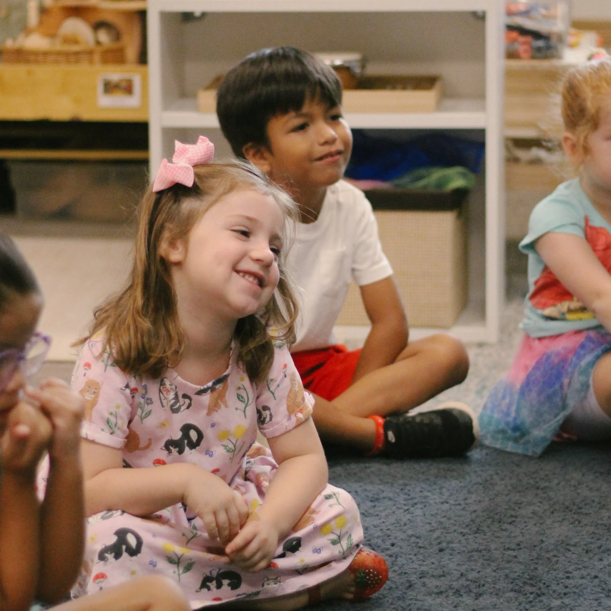 Children sitting on the carpeted floor in a classroom, smiling and laughing.