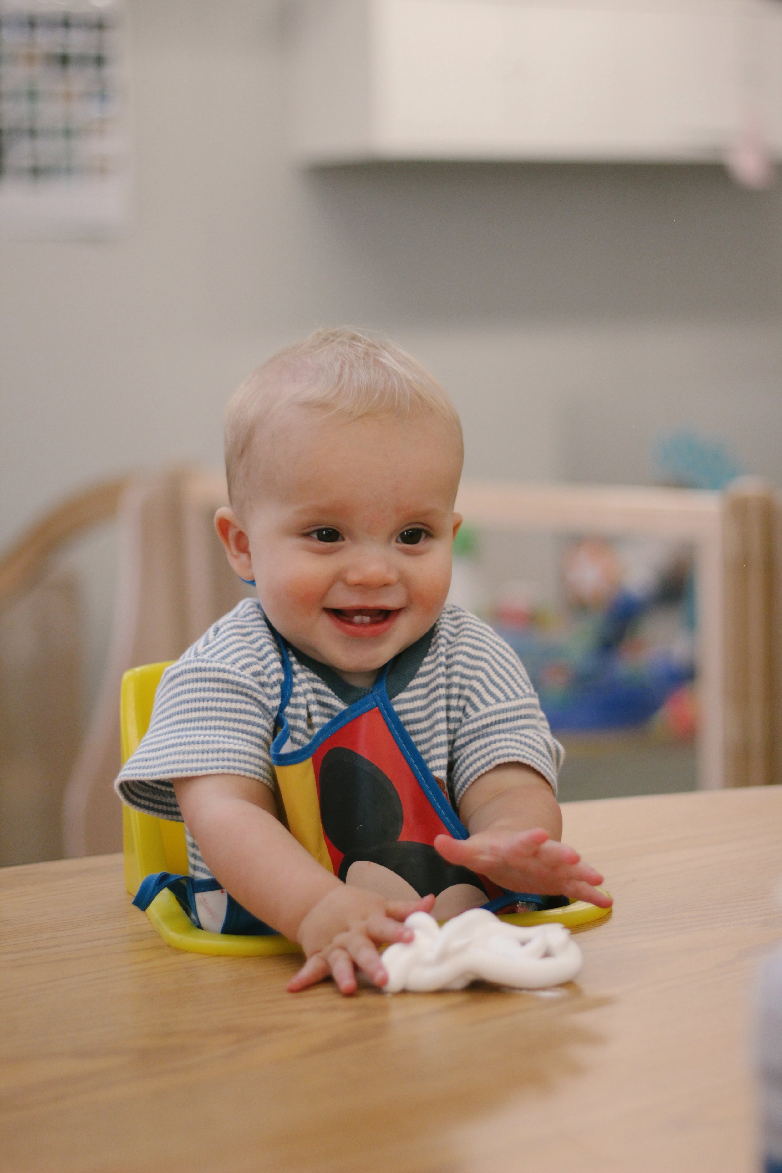 A smiling baby sitting at a wooden table with white slime in front of him.