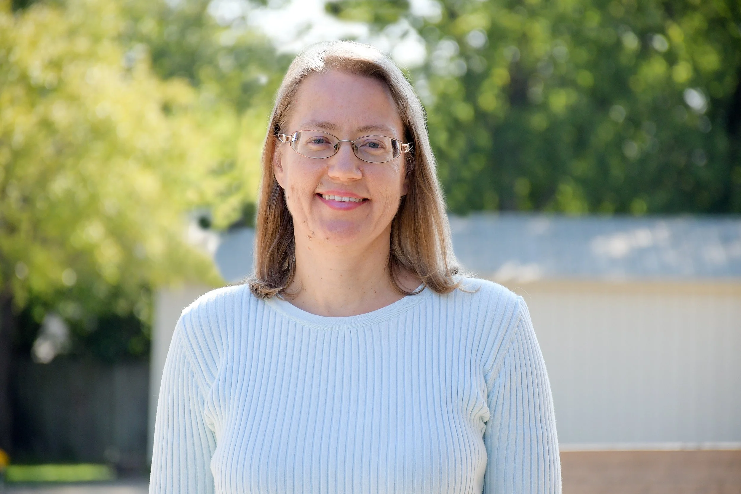 A woman with shoulder-length blonde hair, wearing glasses and a white sweater, smiling outdoors with green trees and a fence in the background.
