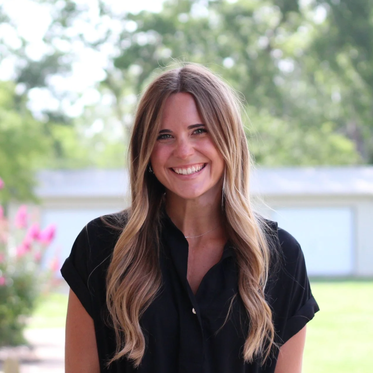 A woman with long, wavy, light brown hair smiling outdoors in a garden with blurred green trees and pink flowers in the background.