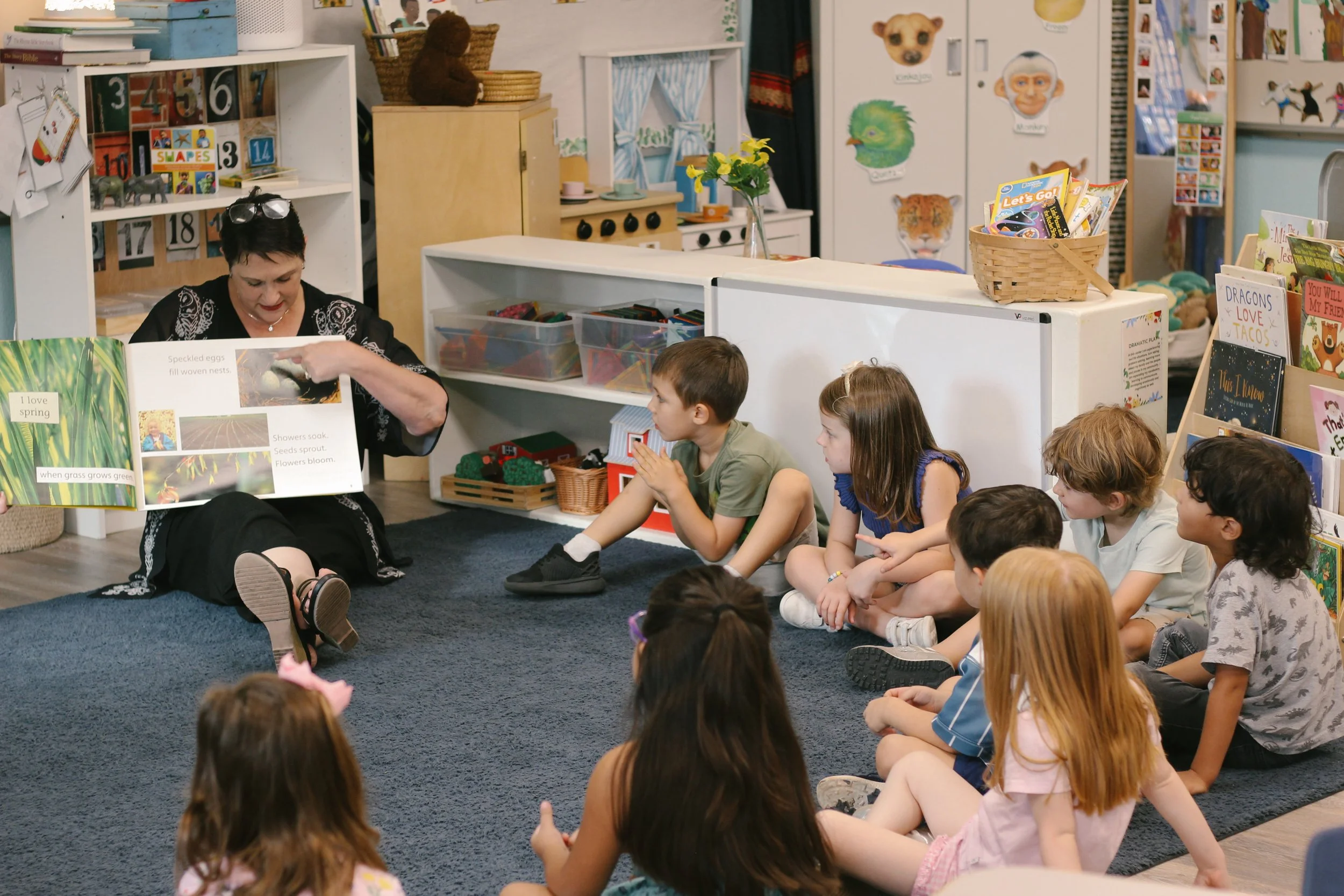 A woman reading a picture book to a group of children seated on a carpet in a classroom.