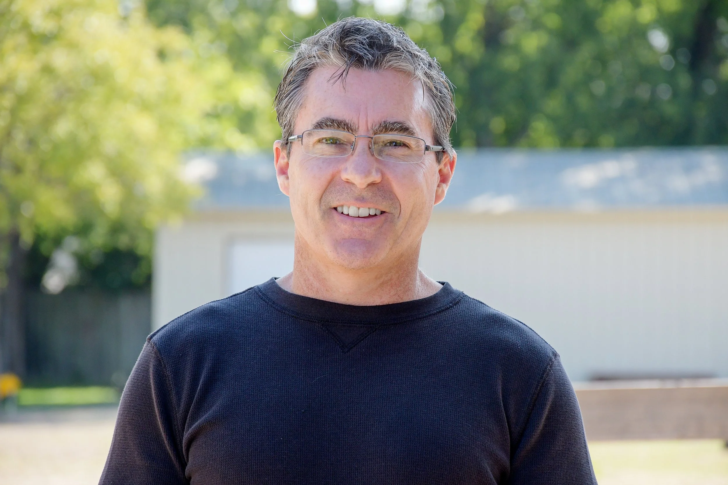 A middle-aged man with glasses, smiling, wearing a black shirt, standing outdoors with trees and a building in the background.