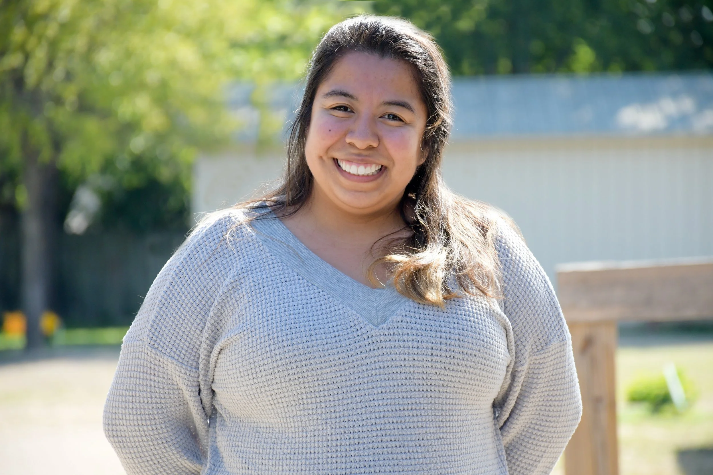 A young woman with long dark hair smiling outdoors on a sunny day, wearing a light gray sweater, with trees and a blue fence in the background.