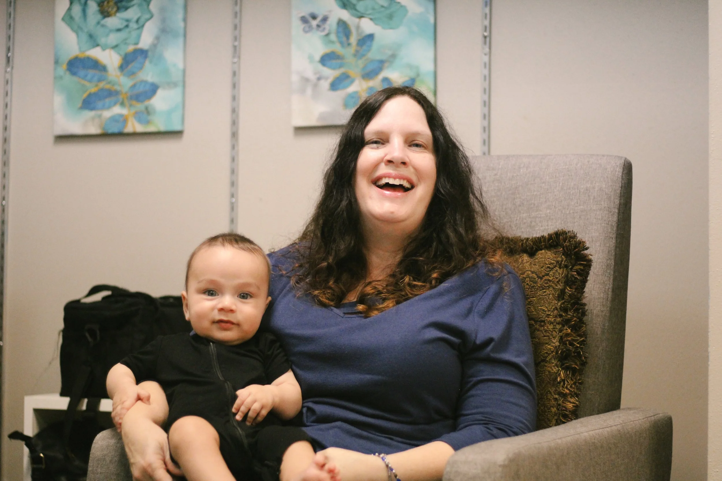 A teacher smiling while sitting in a gray armchair, holding a young baby in a classroom.