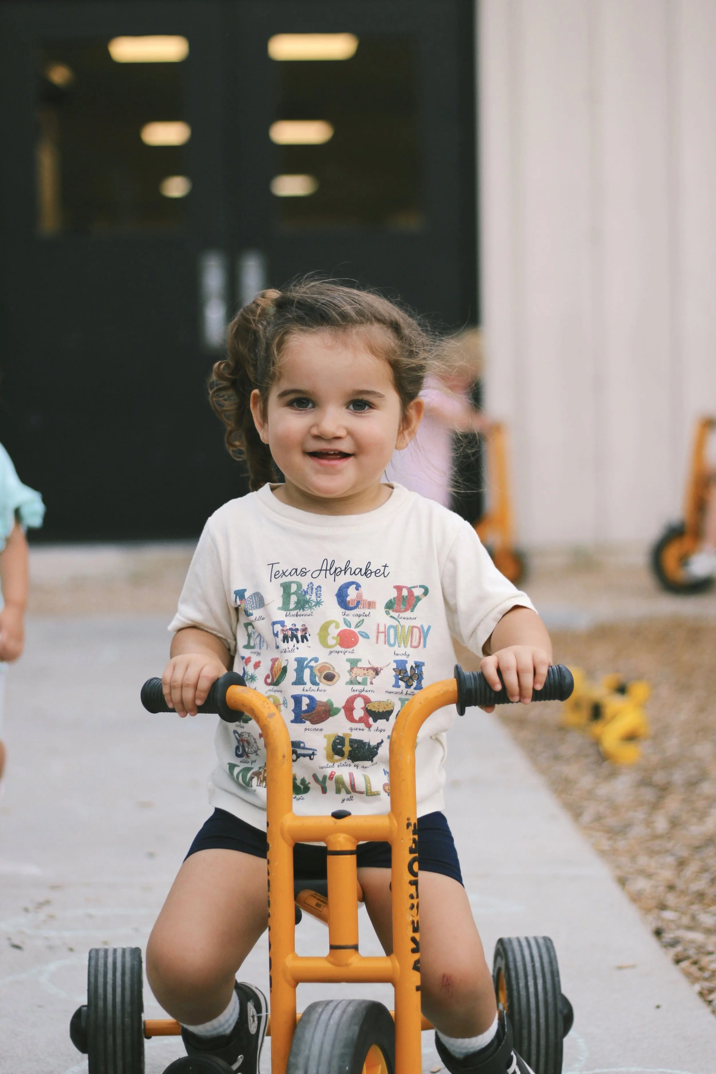 A young girl with curly hair riding an orange tricycle outdoors, smiling at the camera.
