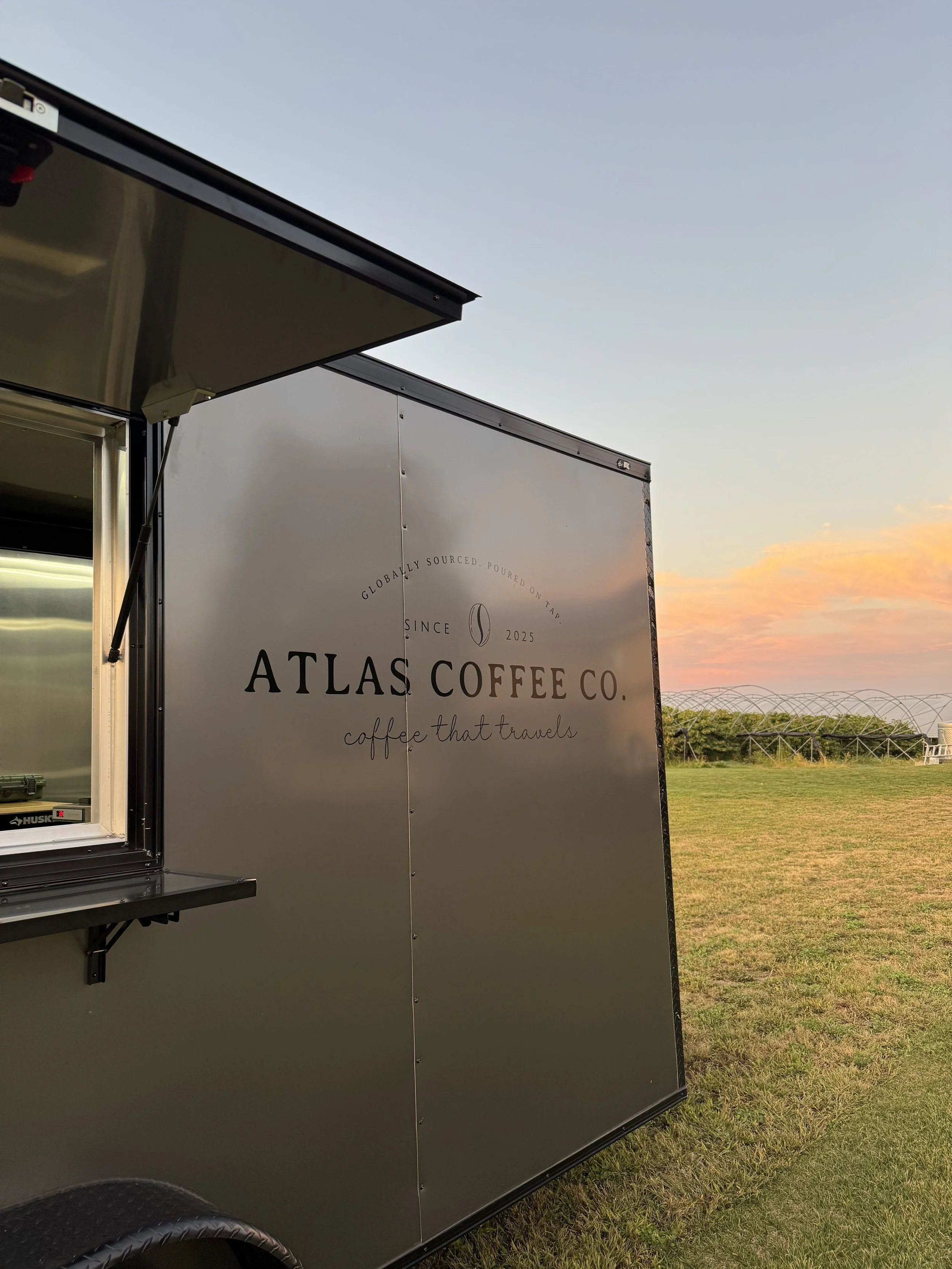A coffee truck with the logo 'Atlas Coffee Co.' parked outdoors during sunset, with open window and grassy field in the background.