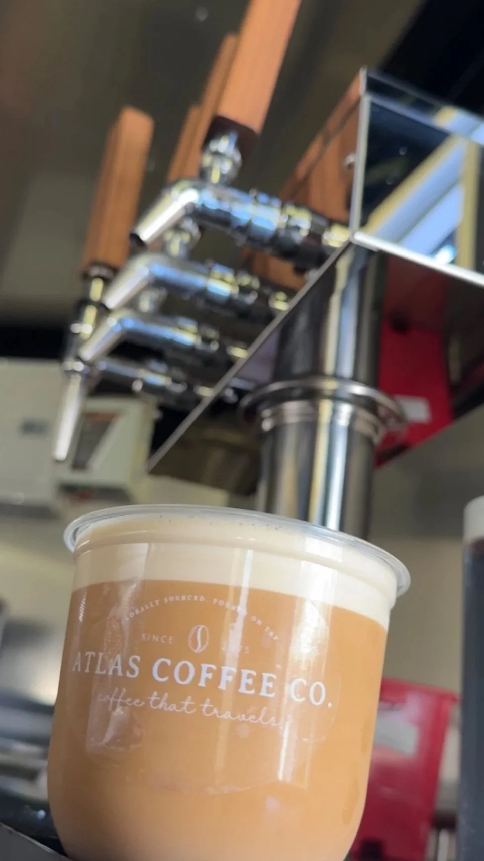 Close-up of a plastic cup with the logo 'Atlas Coffee Co.' on it, filled with a layered coffee drink, placed on a surface in front of a professional espresso machine.