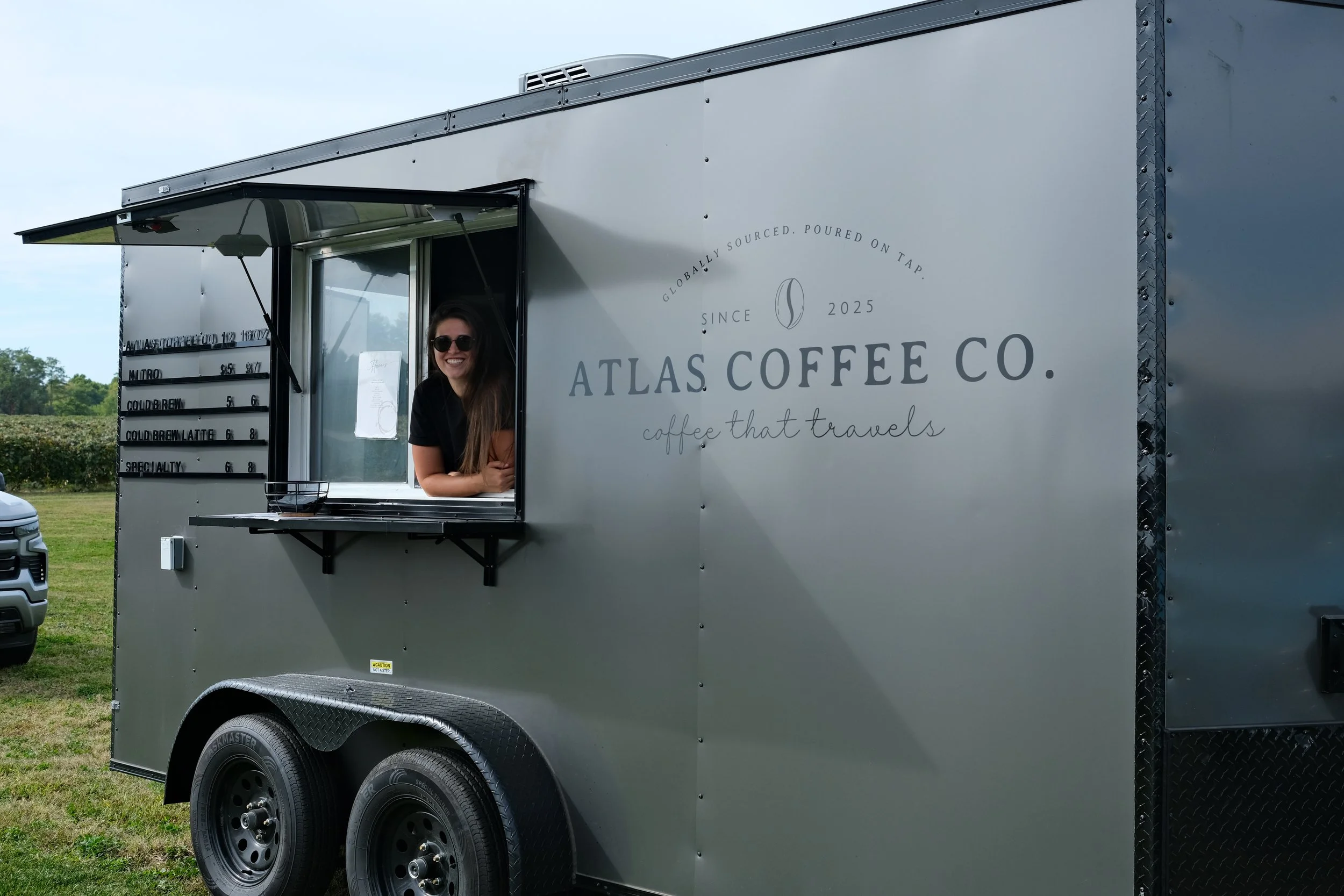 A smiling woman wearing sunglasses is leaning out of a window of a gray coffee trailer with the sign 'Atlas Coffee Co.' on the side. The trailer has part of a menu visible and is parked outdoors on grass.