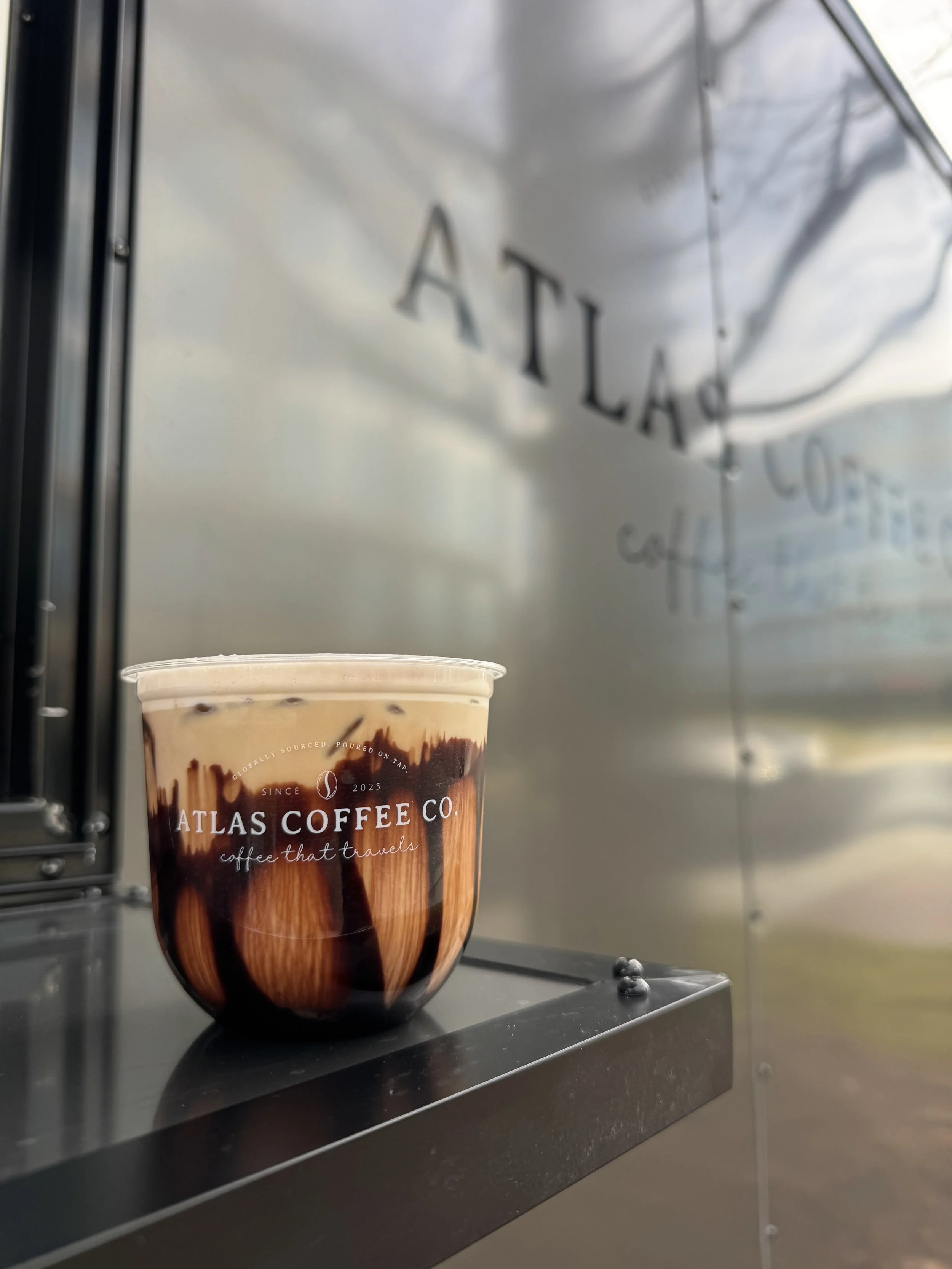 A plastic cup filled with iced coffee topped with whipped cream and drizzled with chocolate syrup, placed on a black surface outside at ATLAS COFFEE CO.