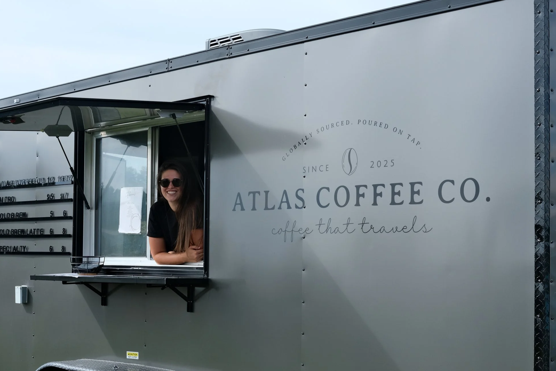 Smiling woman in sunglasses leaning out of a coffee truck window, which has 'Atlas Coffee Co.' branding and slogan 'coffee that travels' on its side