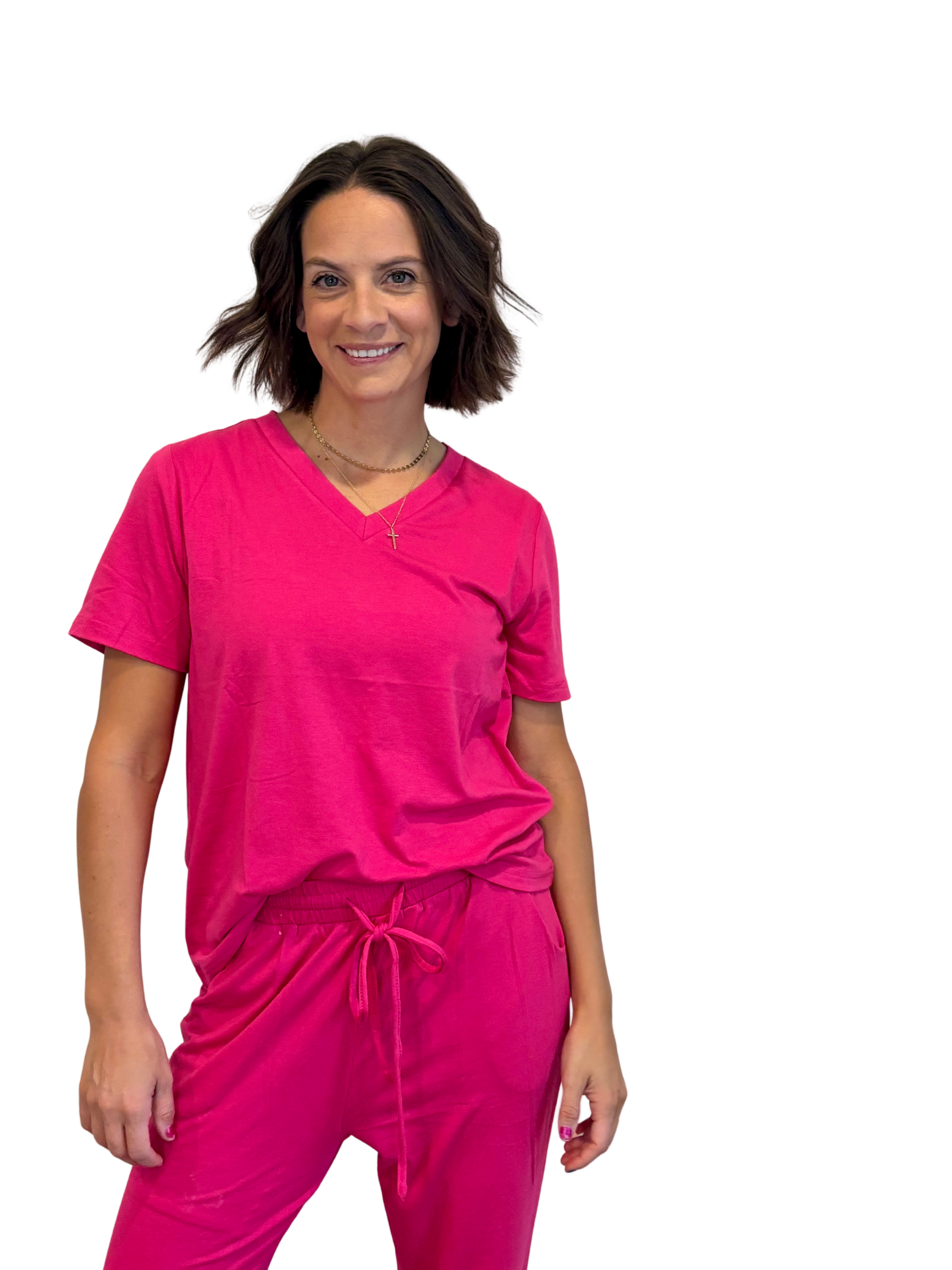 A woman with shoulder-length brown hair smiling, wearing a pink t-shirt and matching pink pants with a drawstring, accessorized with a gold cross necklace and a short gold chain.