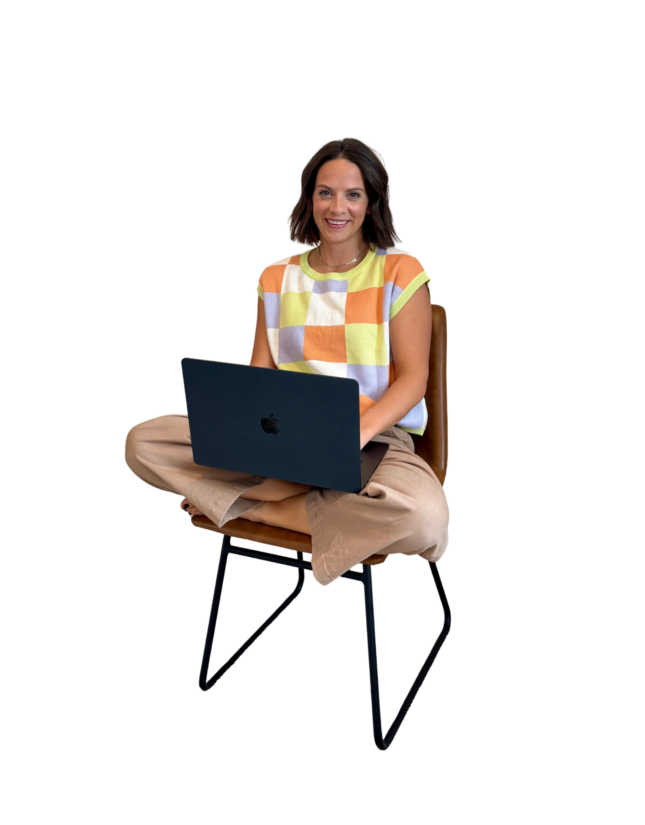A woman sitting cross-legged on a brown chair, smiling and working on a black laptop.