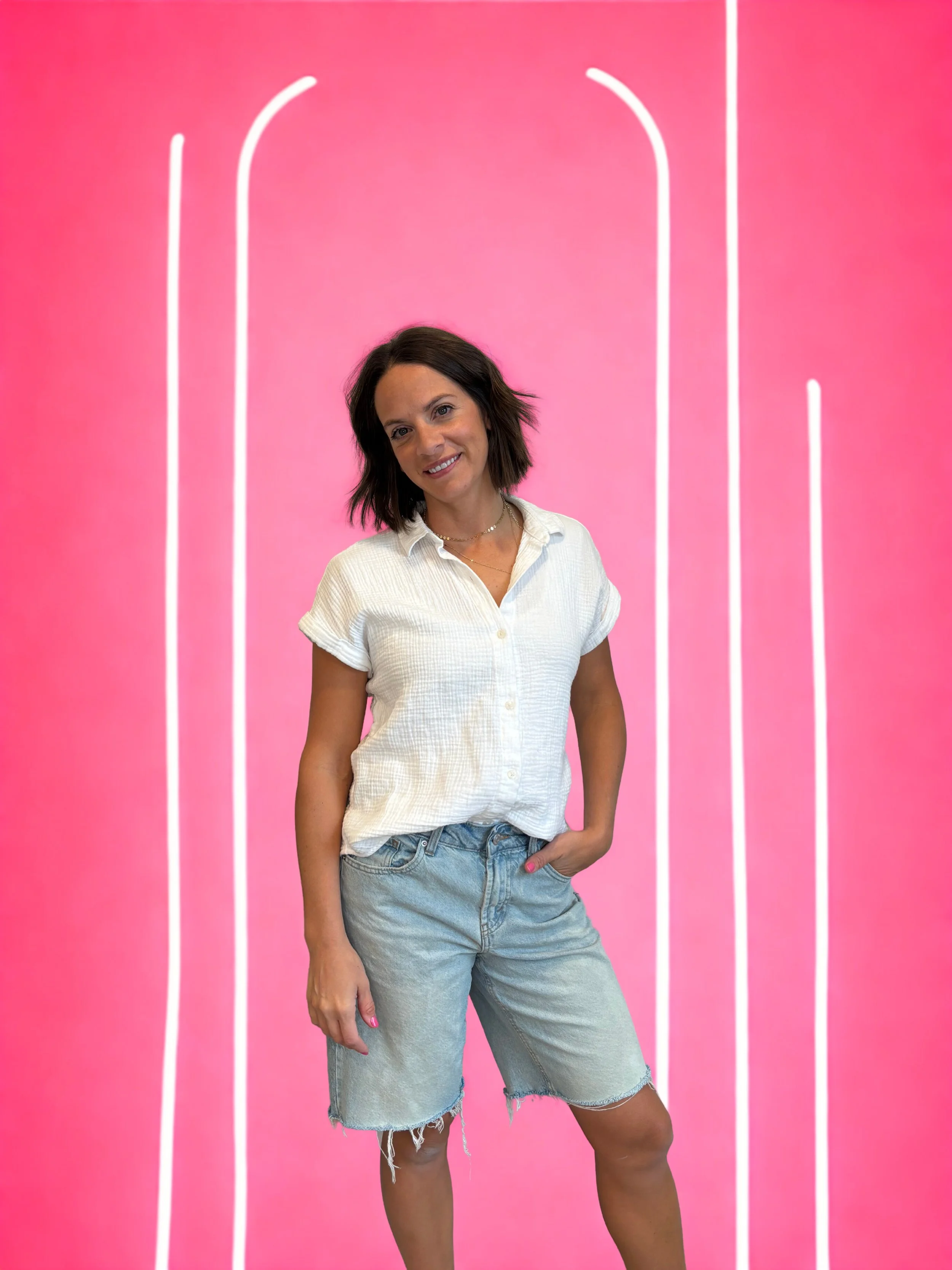 A woman standing in front of a pink background with white decorative lines, wearing a white short-sleeve shirt and denim shorts.