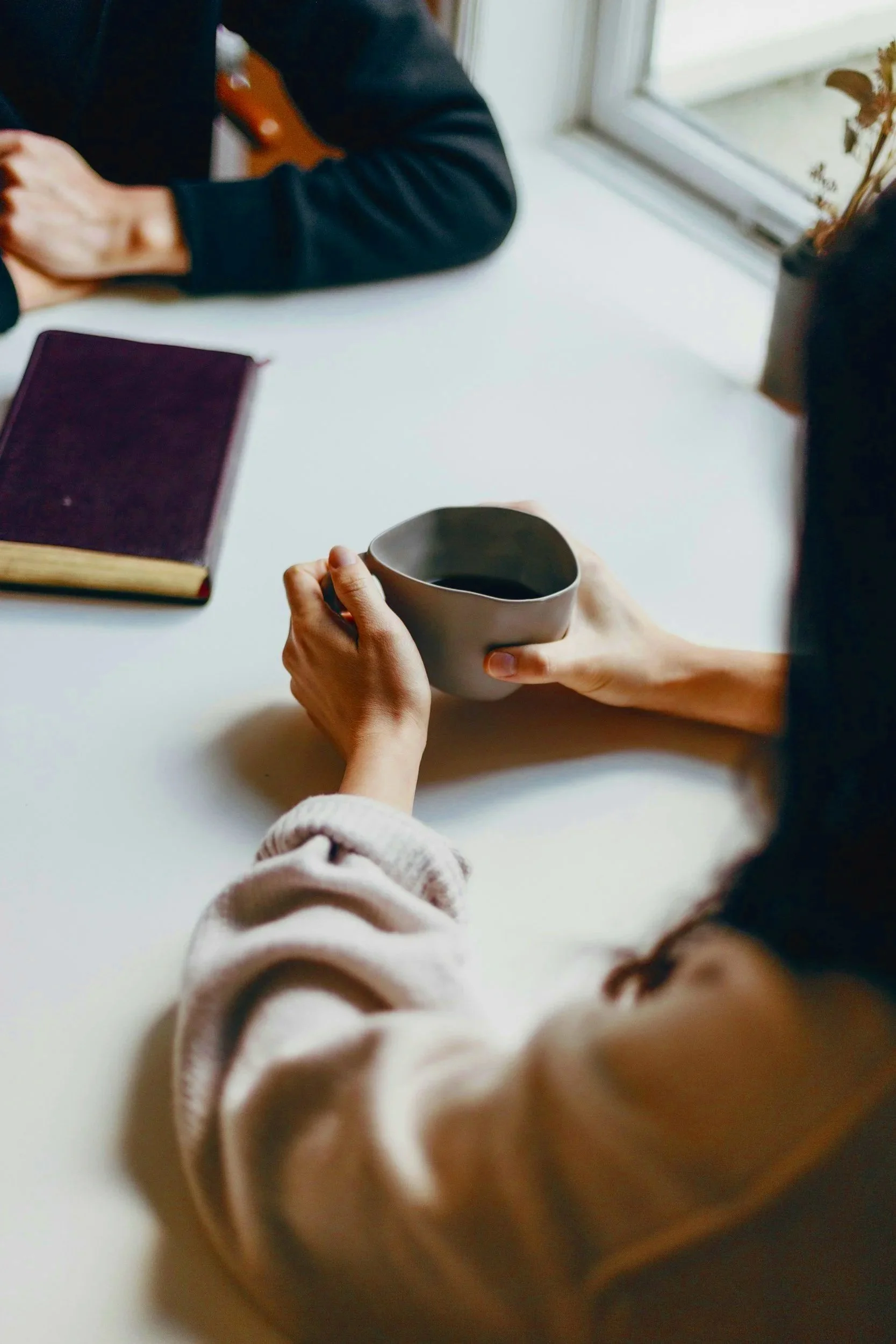 Person holding a ceramic mug with a dark beverage, seated at a table near a window, with another person sitting across, and a closed book with a dark cover on the table.
