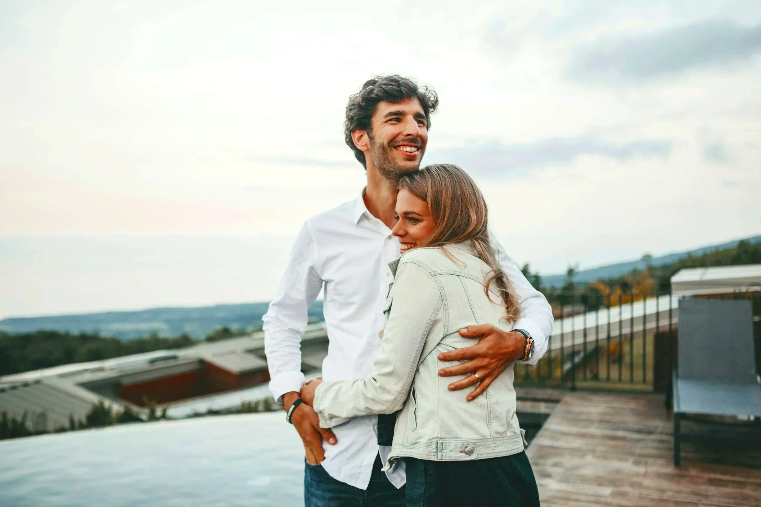 A happy couple hugging outdoors on a rooftop with a scenic landscape in the background, during cloudy weather.