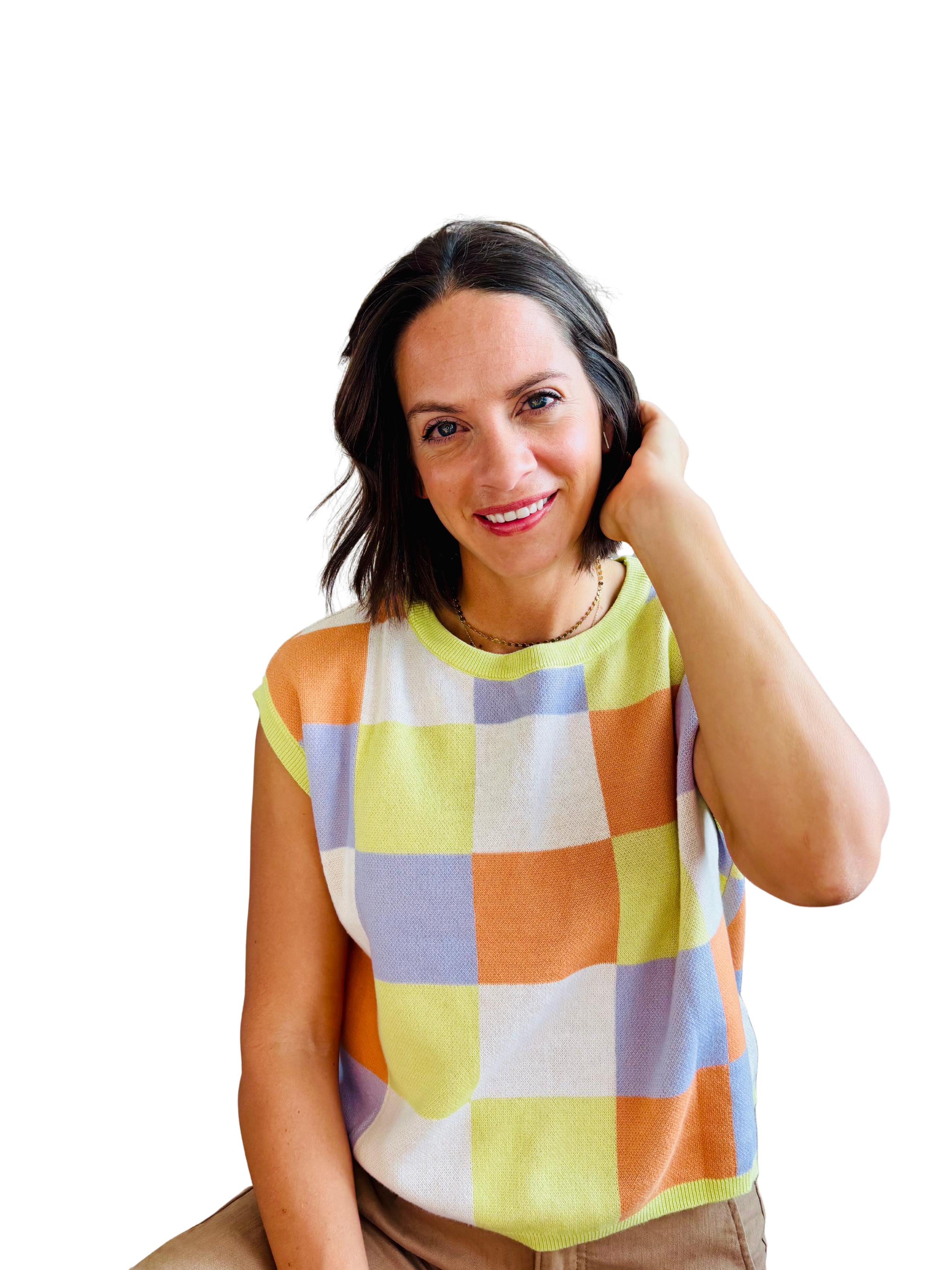 A woman with shoulder-length dark hair, wearing a colorful checkered sleeveless top in shades of yellow, orange, white, and purple, smiling and touching her hair.