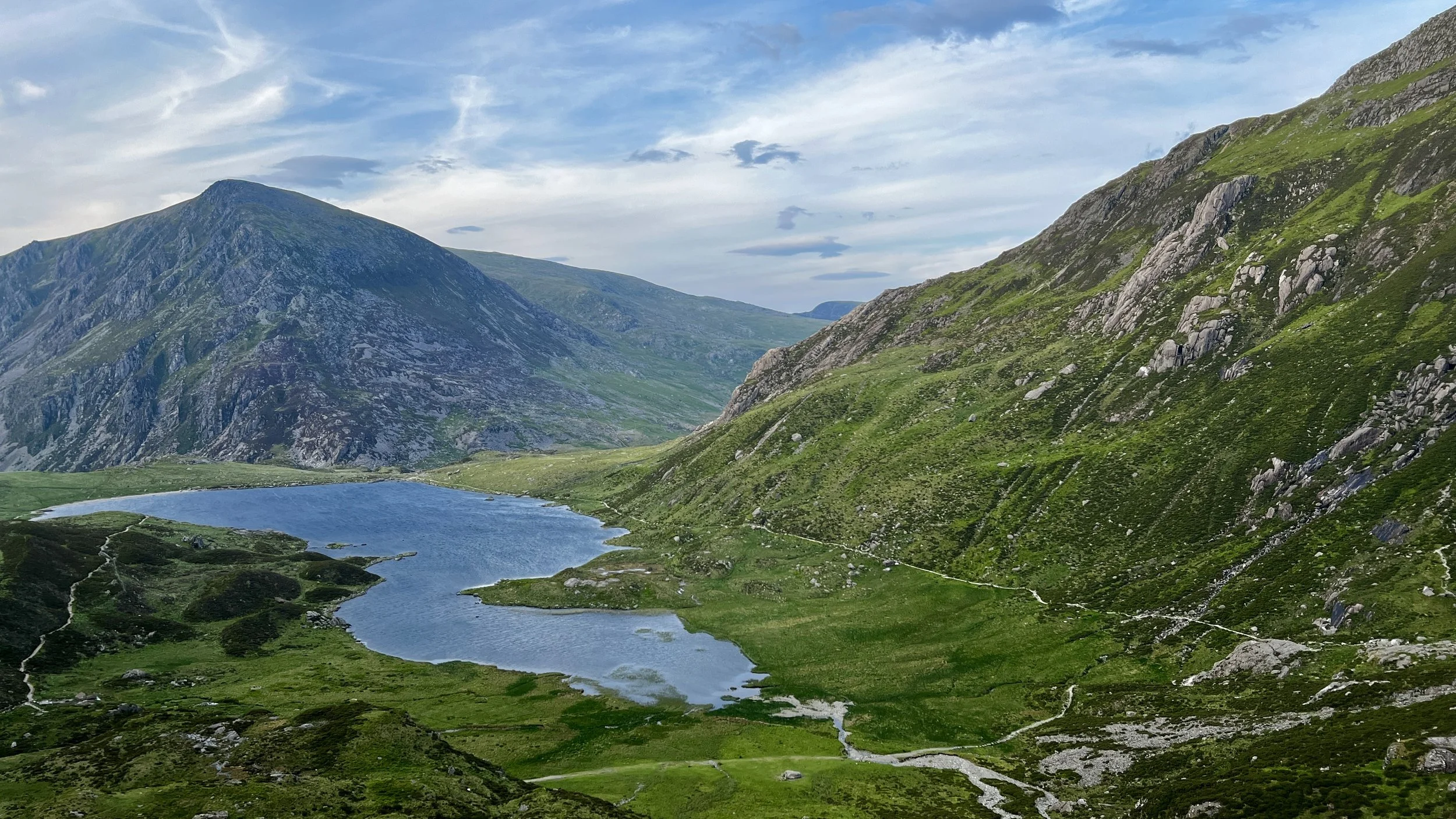 A scenic mountain landscape with green hills and rocky slopes surrounding a blue lake under a partly cloudy sky.