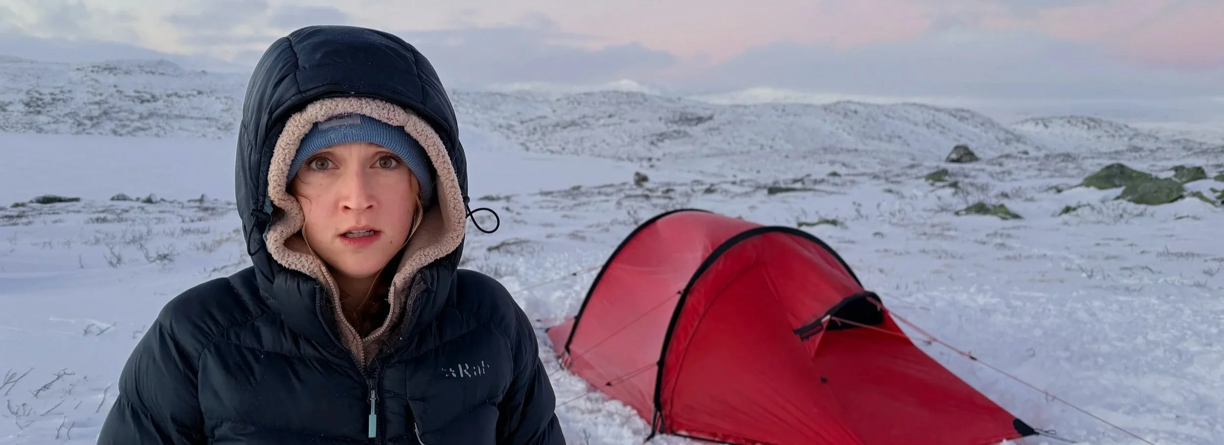 Woman in winter clothing standing in snowy landscape with a red tent behind her.