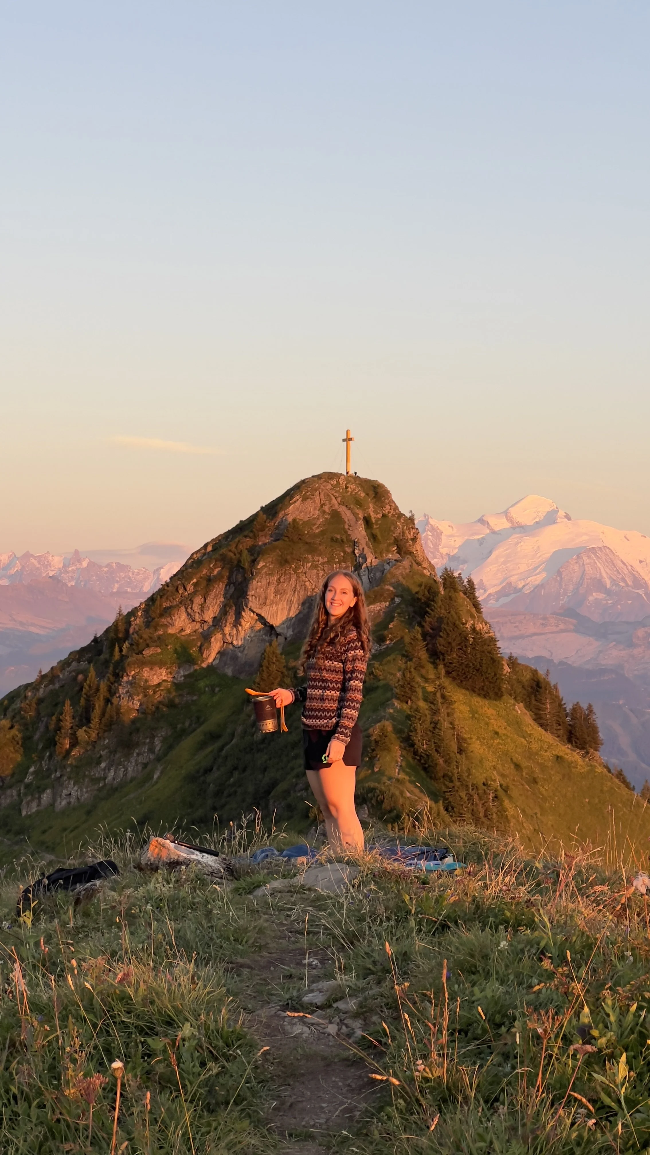 A woman standing on a grassy mountain trail with a mountain landscape and a cross on a peak in the background during sunset, holding a black mug and smiling.