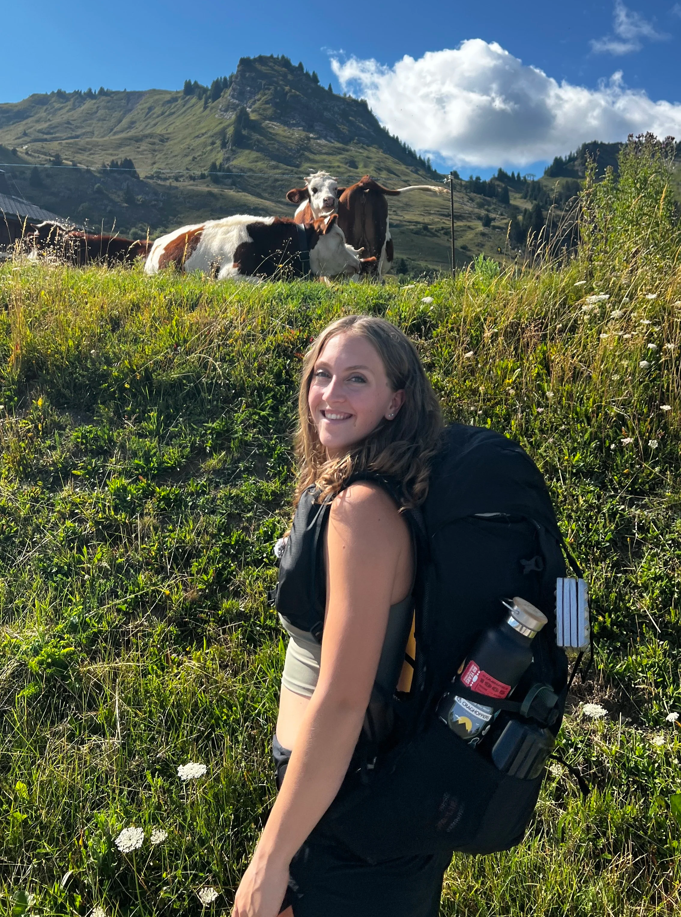 A young woman with long, wavy hair smiling and wearing a black backpack with a water bottle attached, standing on a grassy hillside with cows in the background and a mountain landscape under a partly cloudy sky.