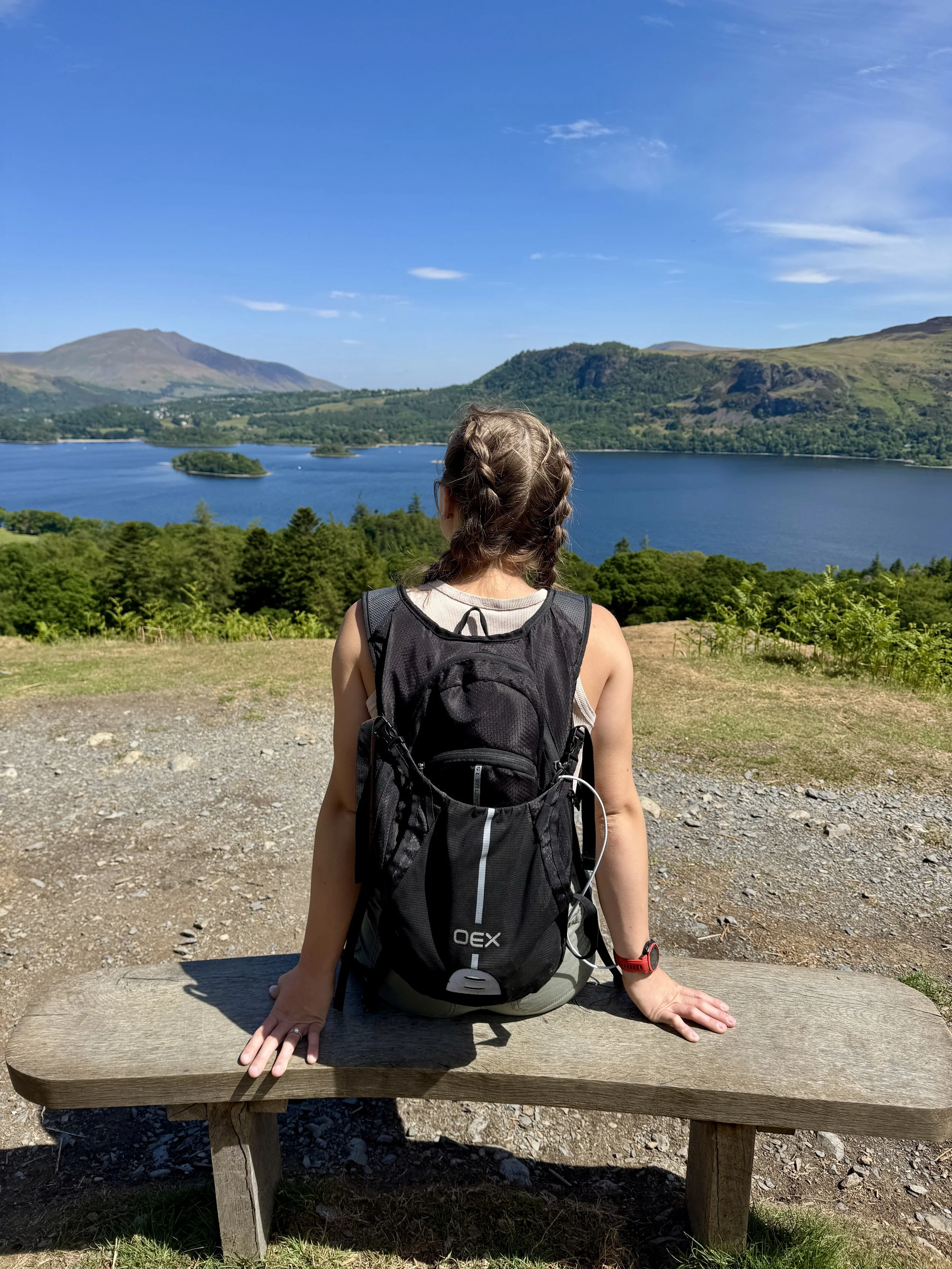 A woman with a backpack sits on a wooden bench overlooking a lake surrounded by green mountains under a clear blue sky.
