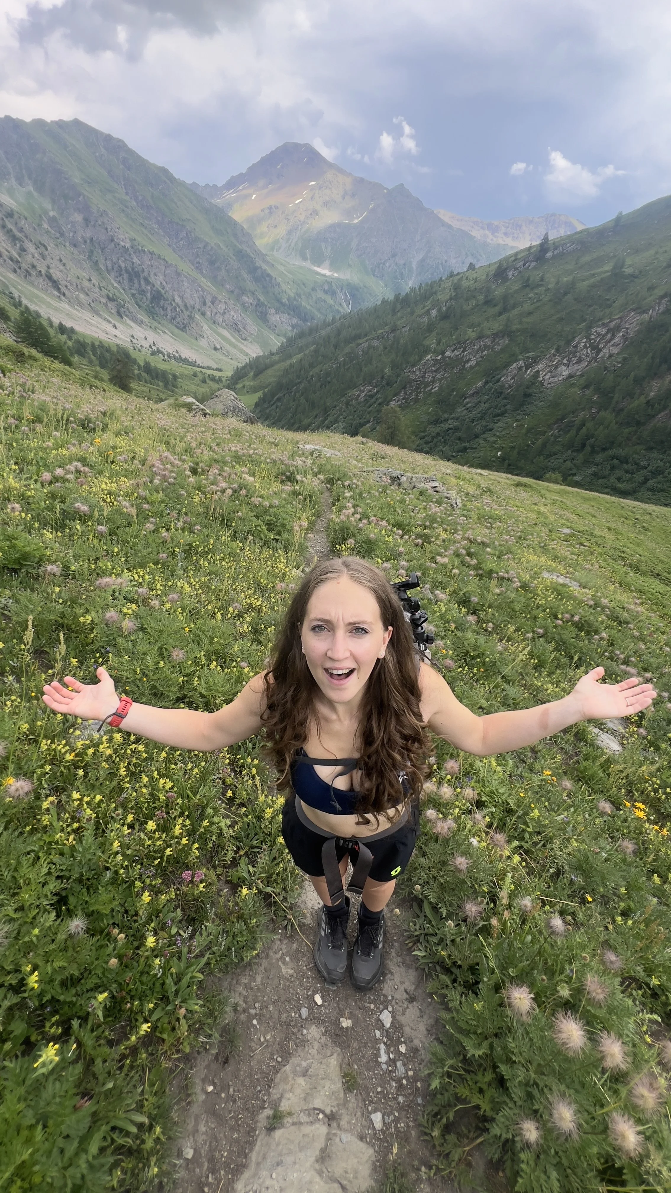 Young woman with long brown hair, wearing a black sports bra, shorts, and hiking boots, standing on a trail in a mountainous area with green hills and rocky peaks in the background, surrounded by wildflowers.