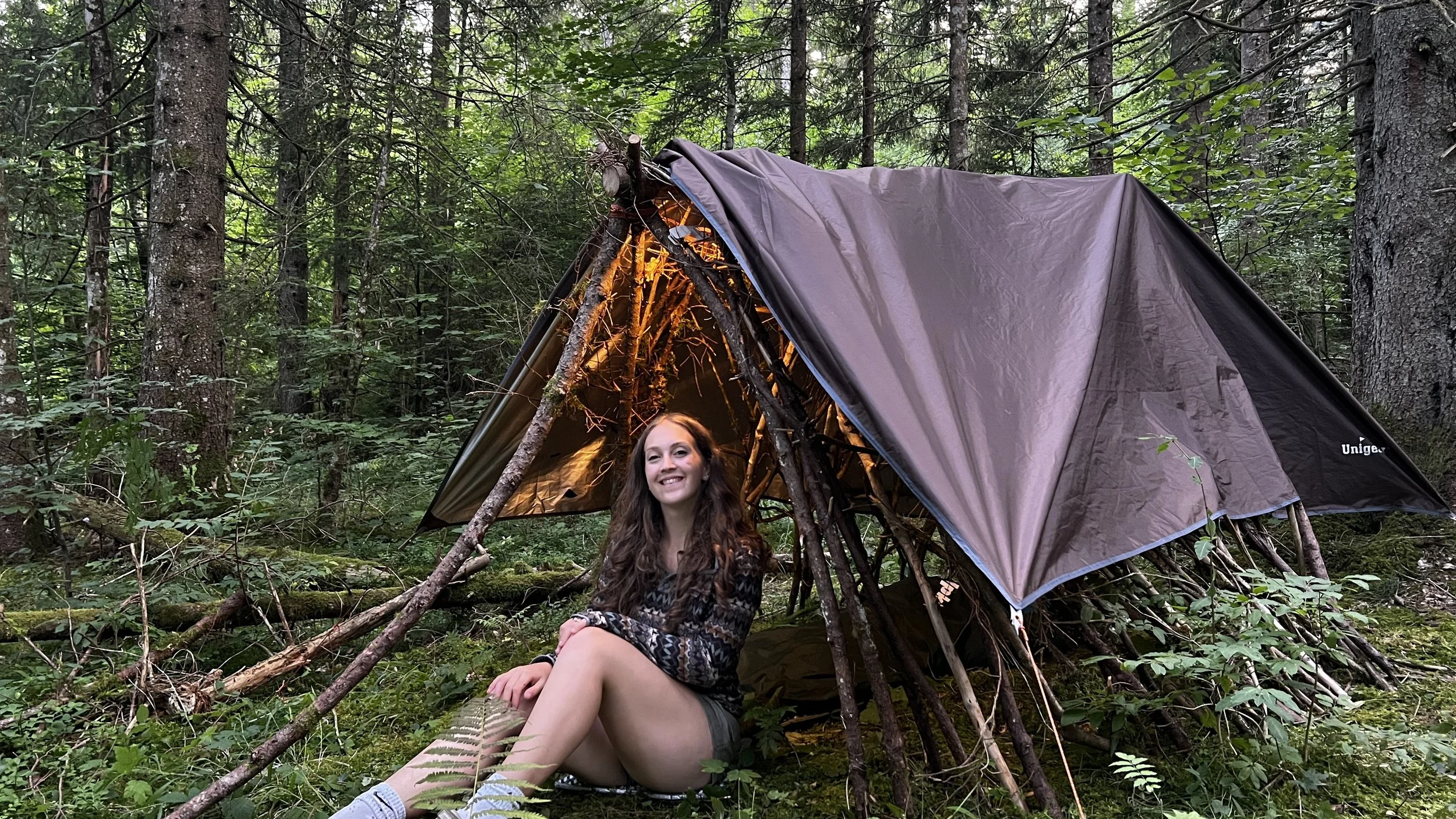 Woman sitting in front of a makeshift shelter in a dense forest, smiling at the camera.