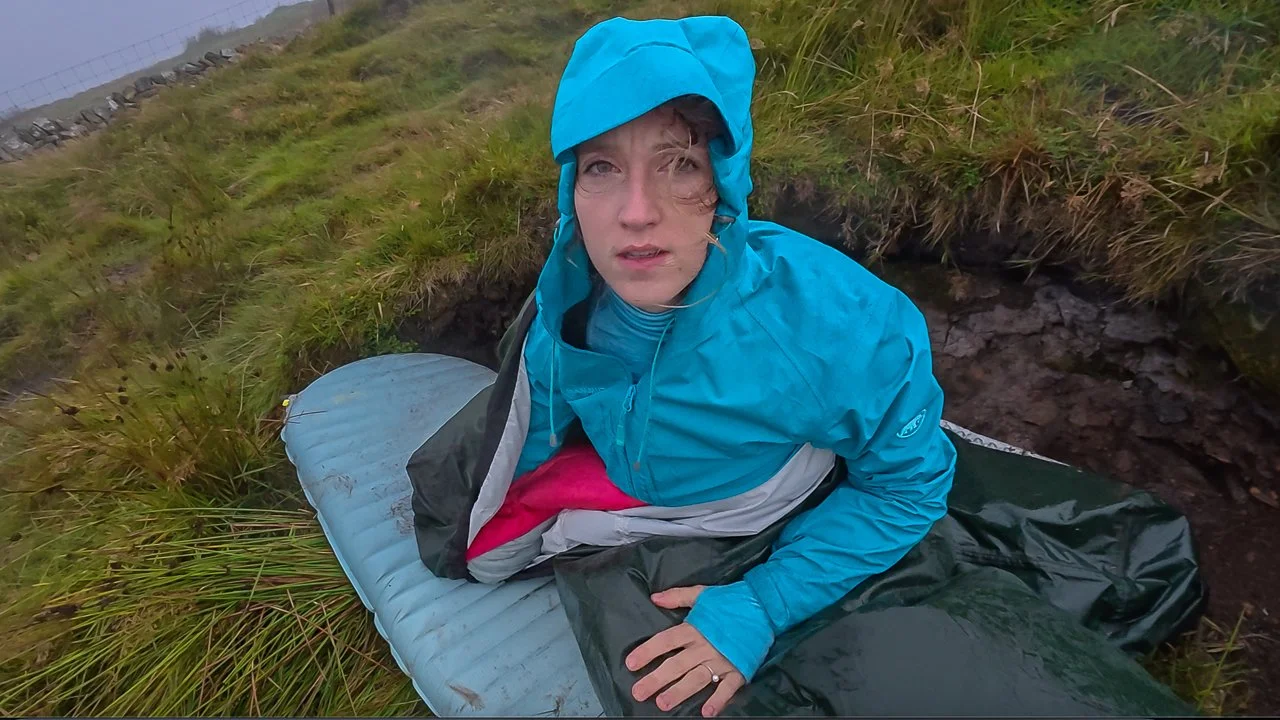 Person in a blue rain jacket and hood sitting on a sleeping pad outdoors near a grassy area during rainy weather.