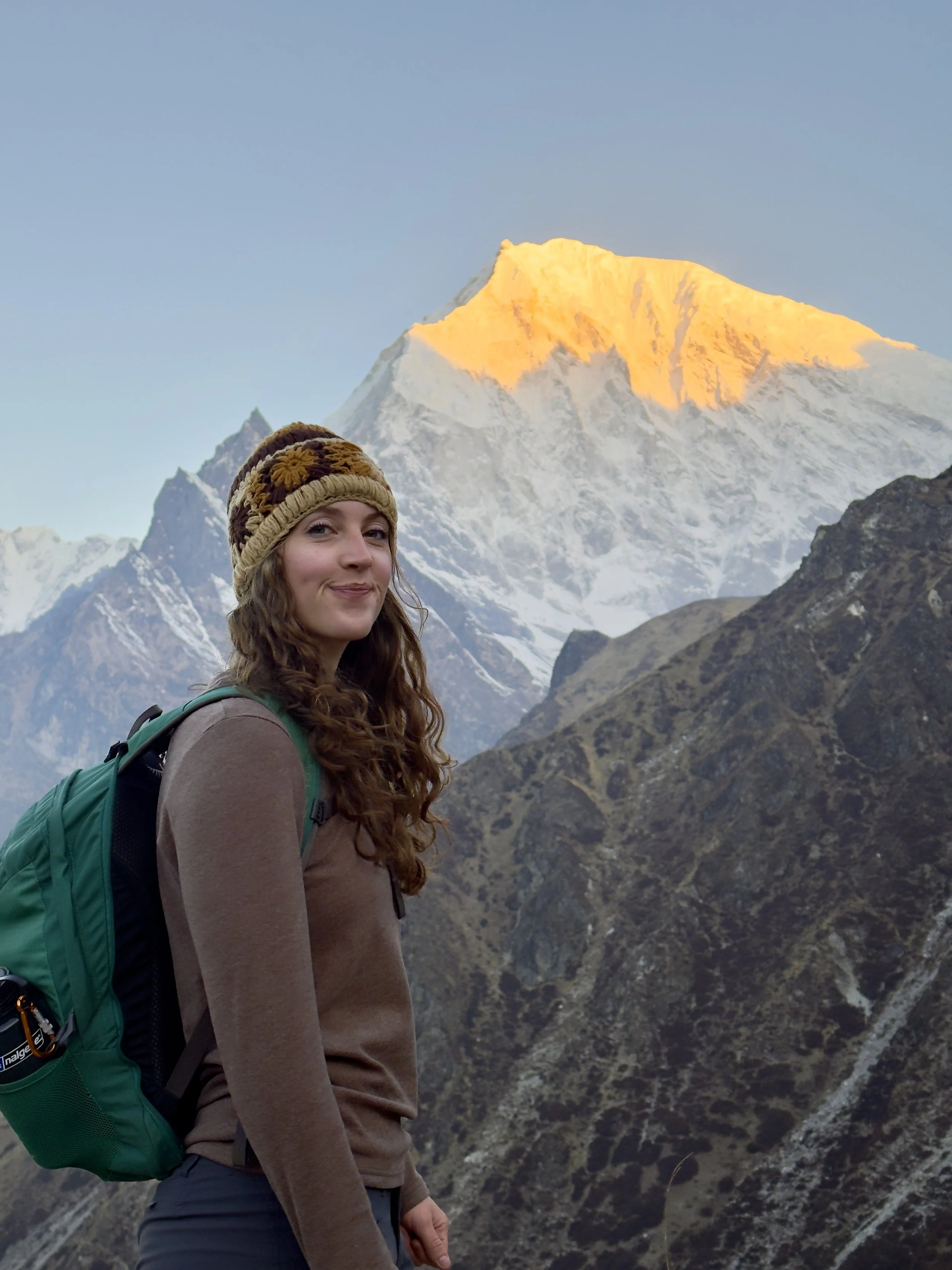 A woman with long curly hair wearing a brown hat, a brown jacket, and a green backpack, standing in front of snow-capped mountains with the peak illuminated by sunlight.