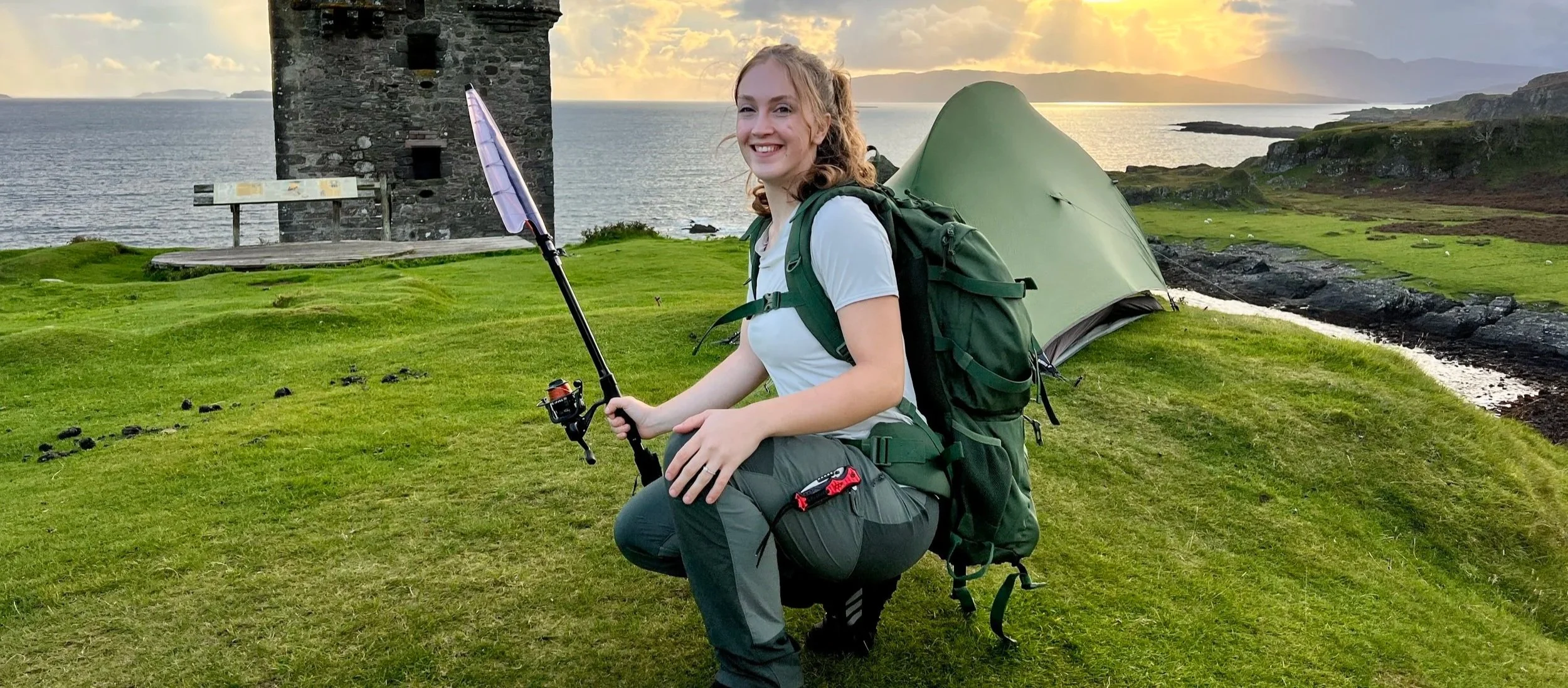 A woman with curly hair and a white t-shirt kneeling on grass, smiling, holding a fishing rod, with a green camping tent and historic stone tower in the background, near a body of water at sunset.