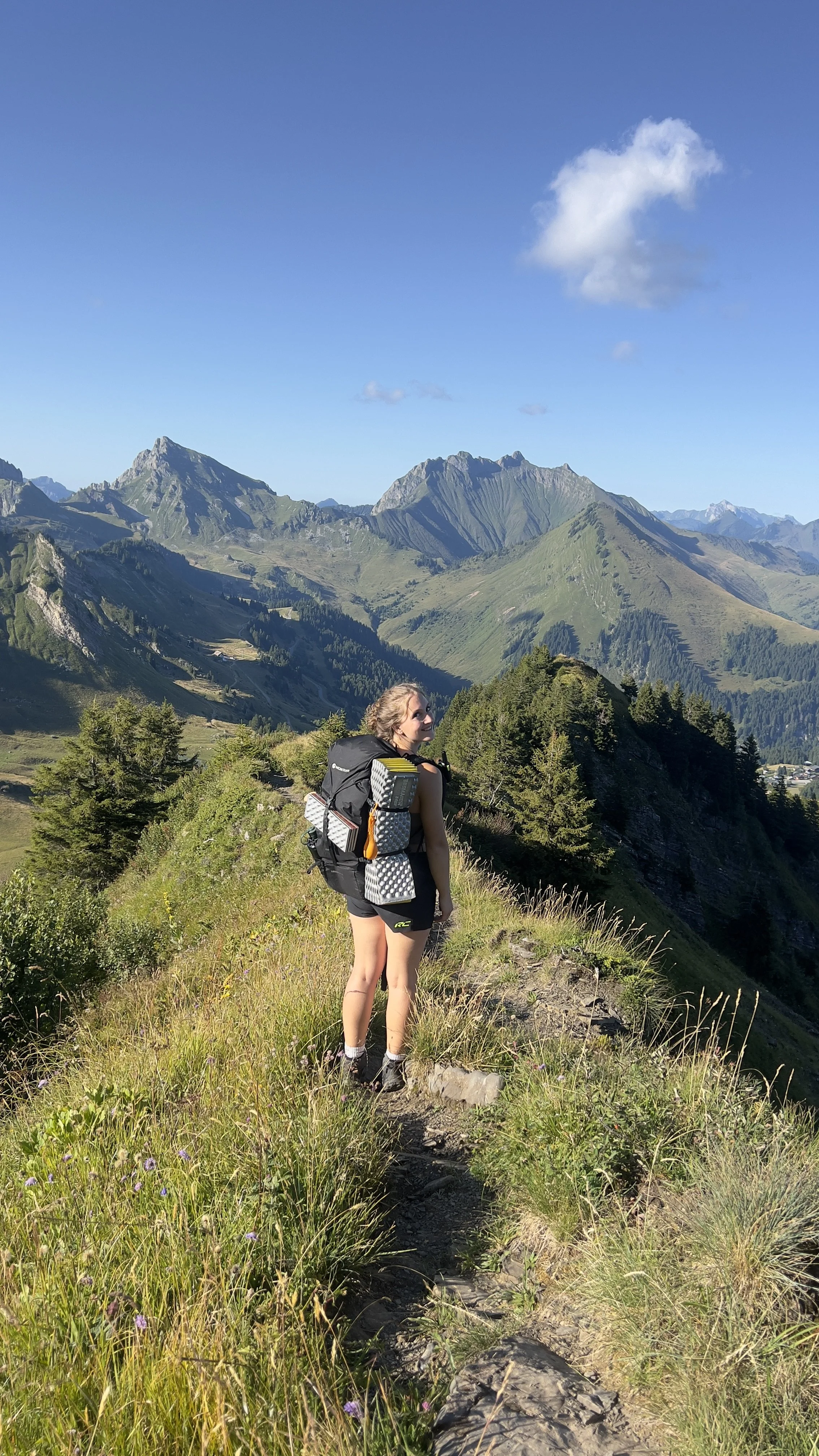 A woman with a backpack hiking along a mountain trail surrounded by grassy hills and trees, with towering mountains and a blue sky in the background.