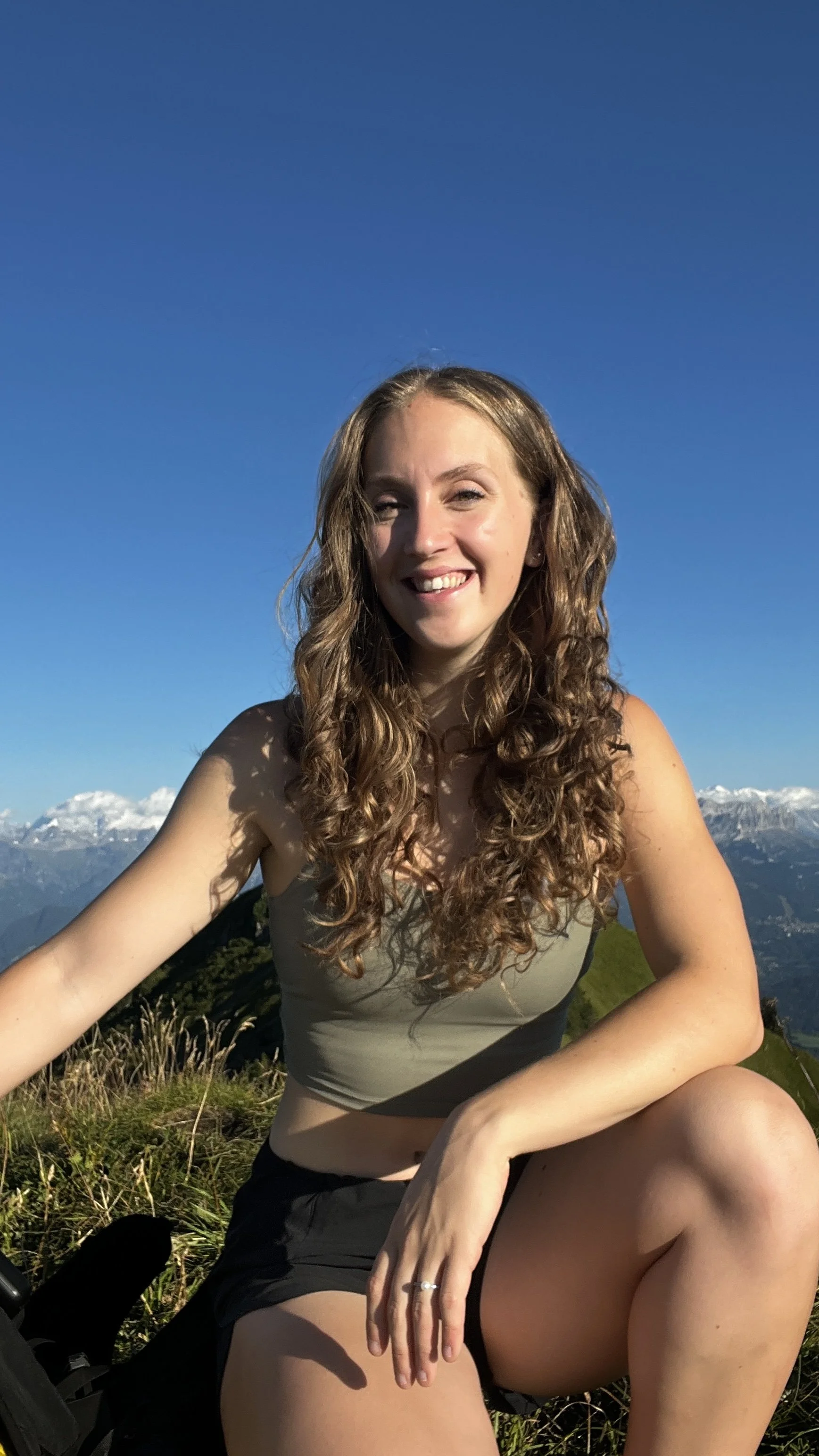 A woman with long curly hair smiling while sitting on a grassy hilltop with mountains and snow in the background under a clear blue sky.