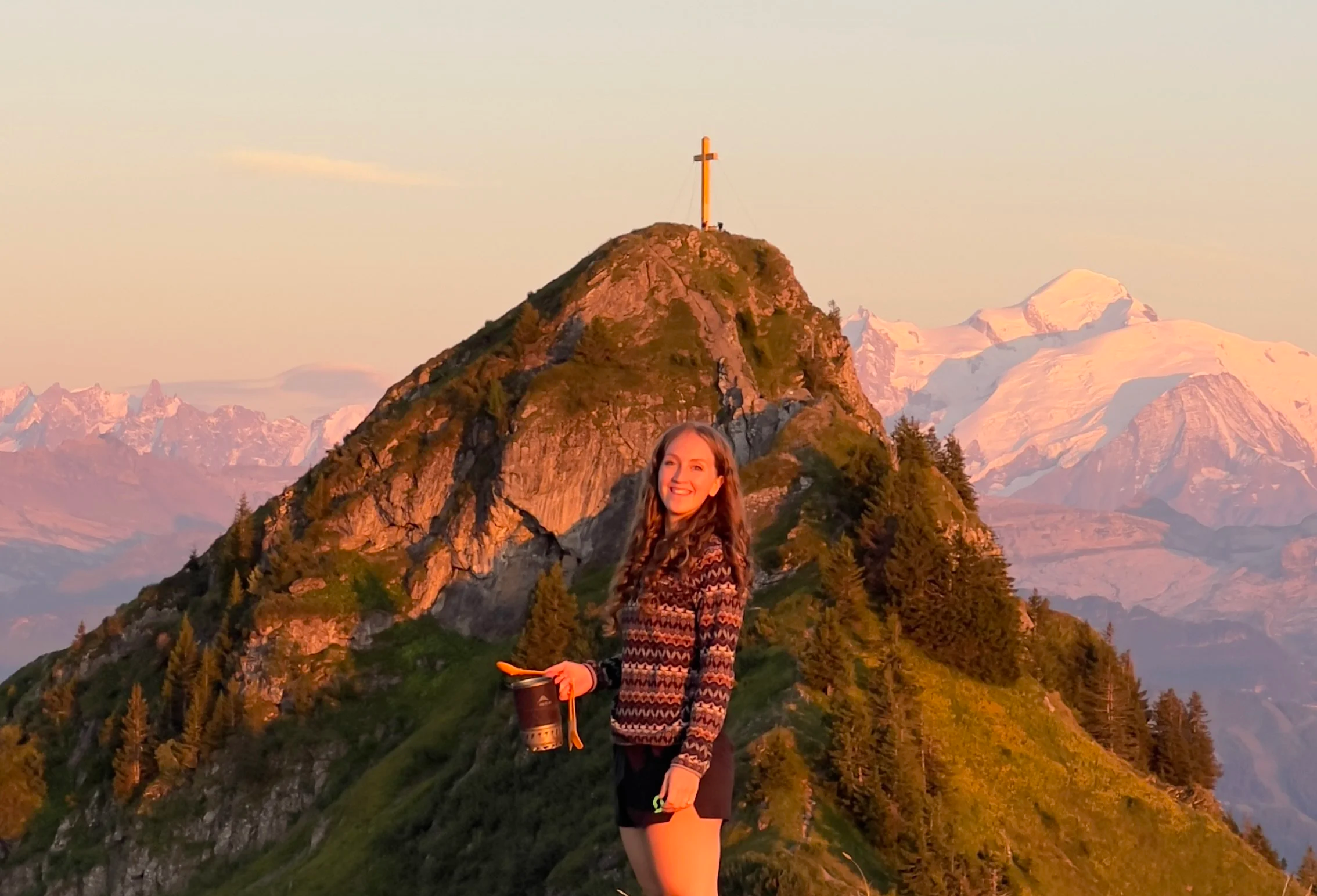 Young woman smiling and holding a mug standing on a mountain trail at sunset, with a mountain with a cross at the summit and snow-capped peaks in the background.