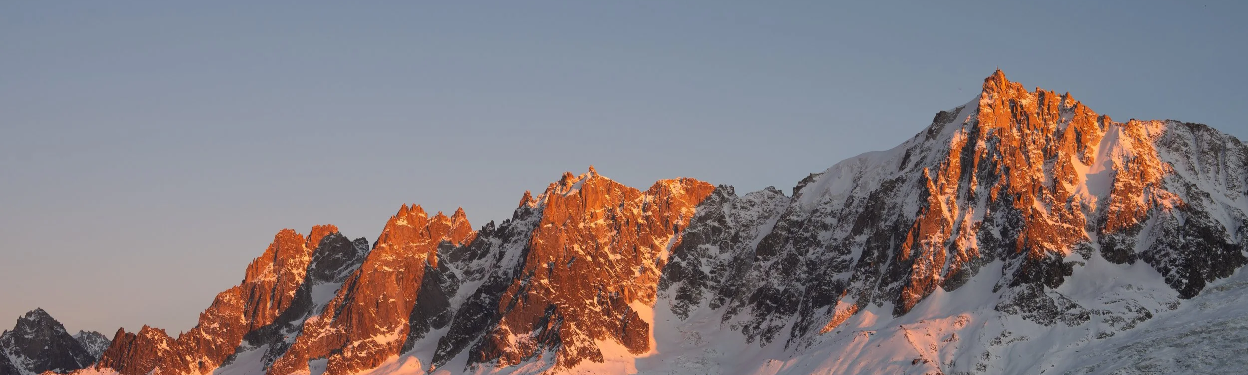 Snow-capped mountain peaks illuminated by orange sunset light under a clear sky.