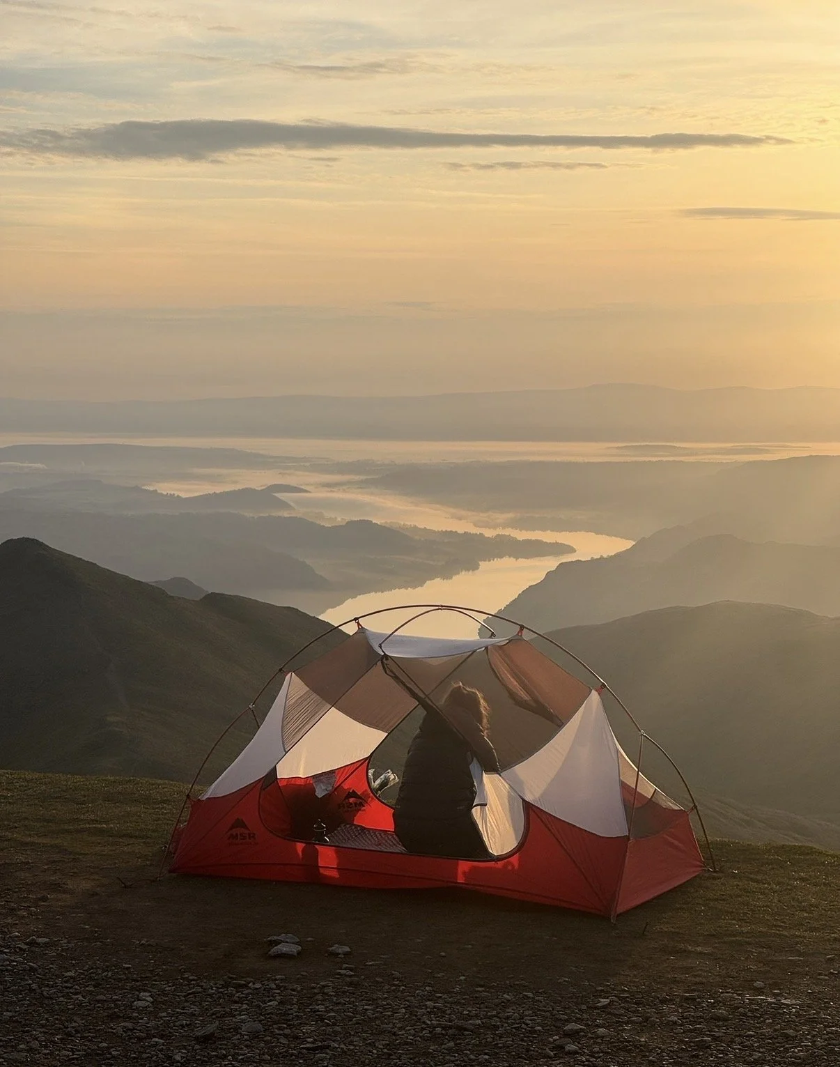 A person sitting inside a red tent on a mountaintop during sunrise, overlooking a landscape with hills, a river, and a distant body of water.
