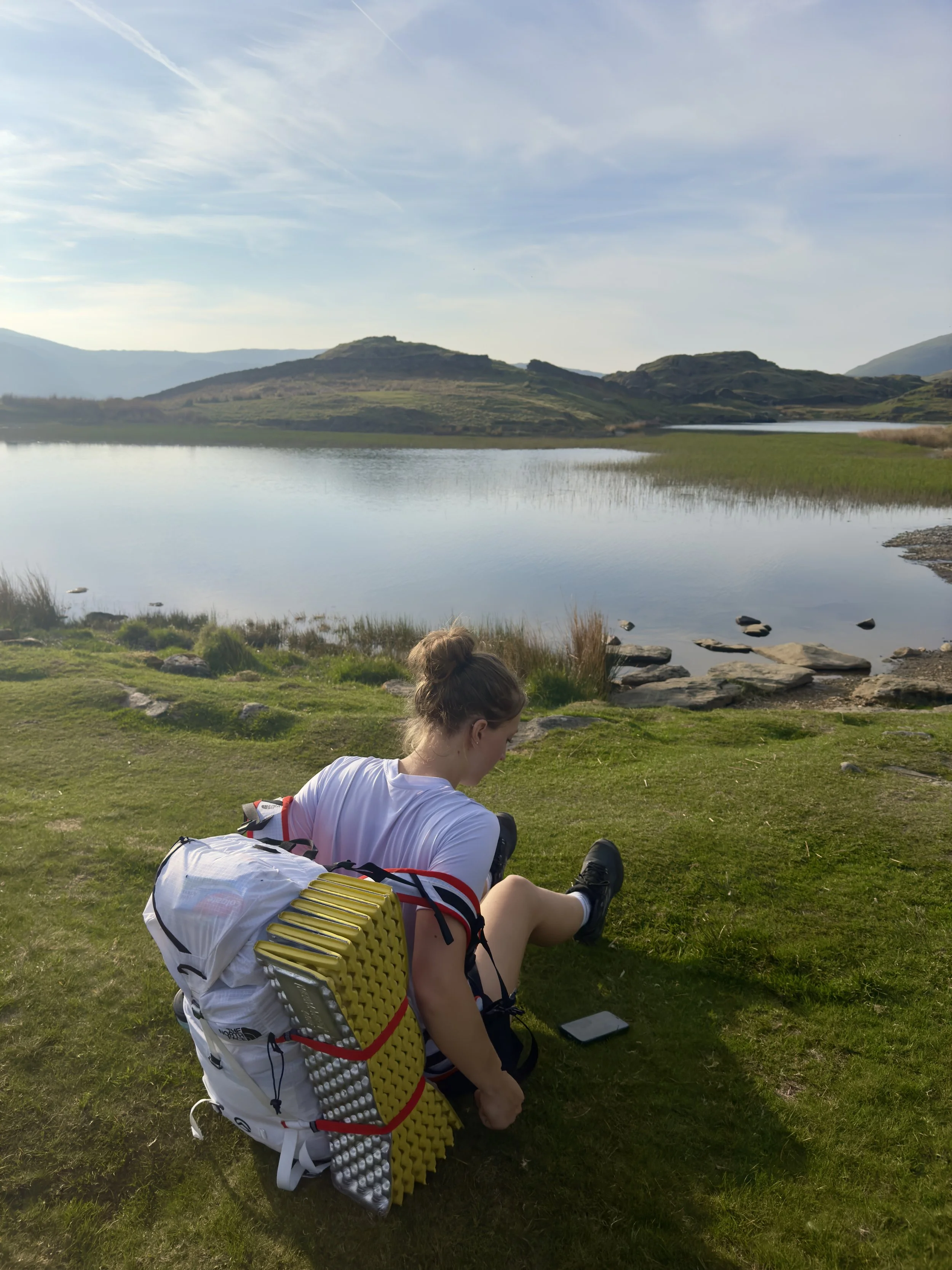 A person relaxing on a grassy lakeshore with a backpack, surrounded by mountains and a calm lake under a partly cloudy sky.