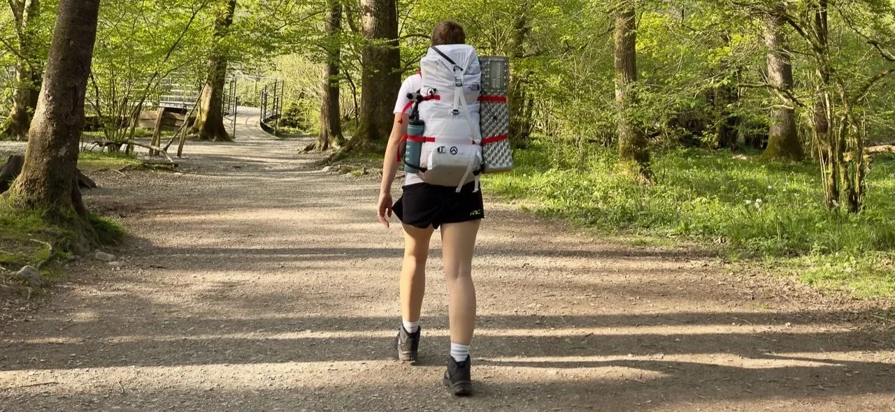 Person with a large backpack hiking on a dirt trail through a forest in spring.