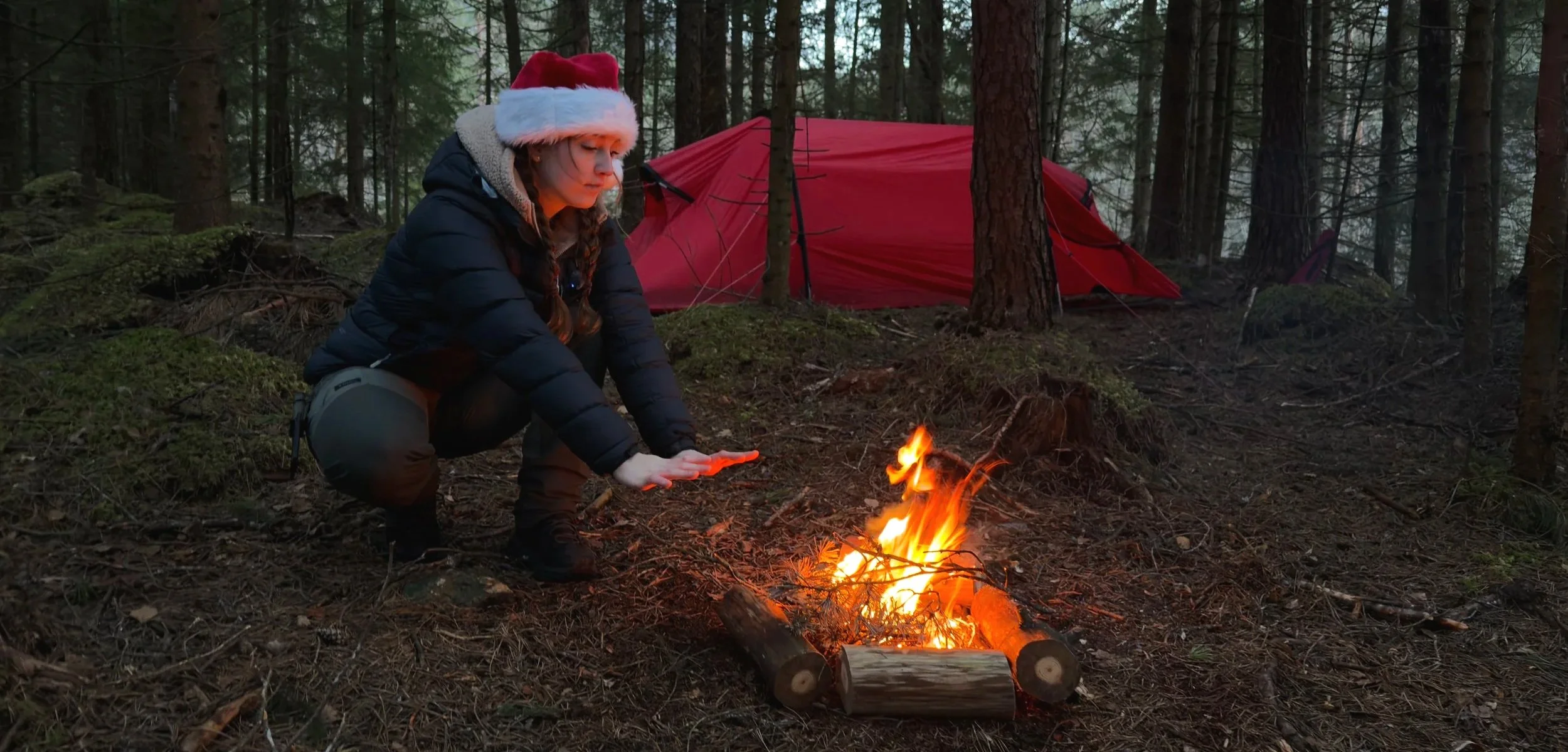 Girl wearing a Santa hat and black jacket squats near a campfire in a forest, with a red tent in the background.